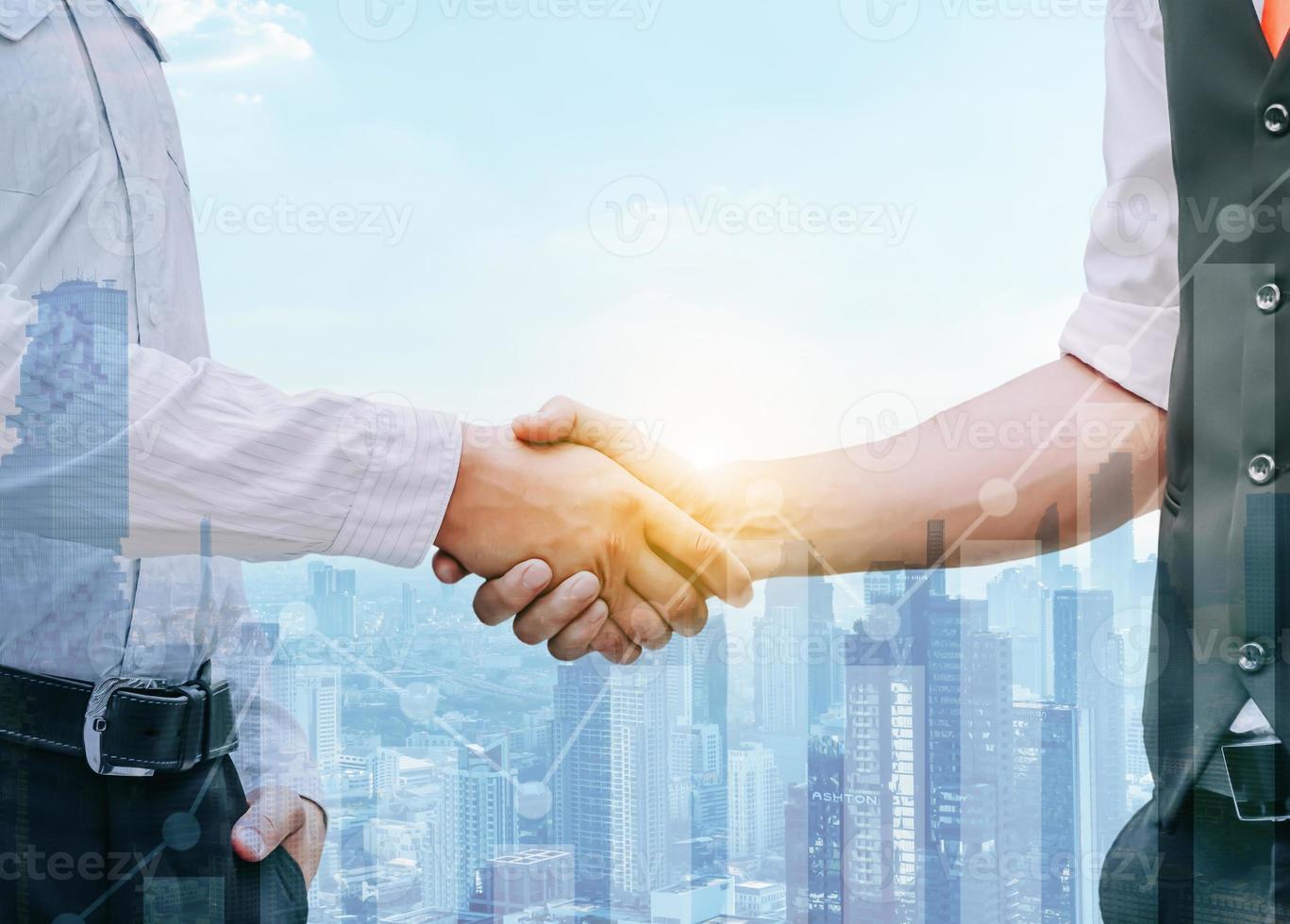 Close up of two businessmen shaking hands against a sunset cityscape. There are glowing graphs in the foreground, Teamwork concept, Concept of stock market deal, Double exposure photo