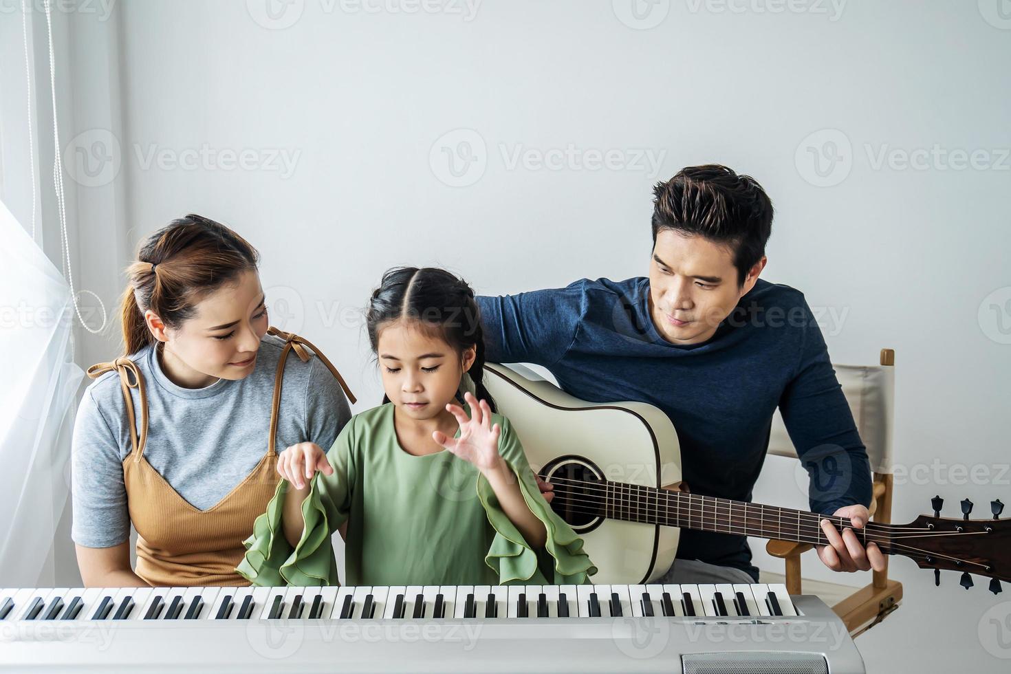 feliz pequeña hija asiática tocando el piano con madre y padre tocando la guitarra en casa, madre enseñando a su hija a tocar el piano, tocan y cantan canciones. Ellos se están divirtiendo. foto