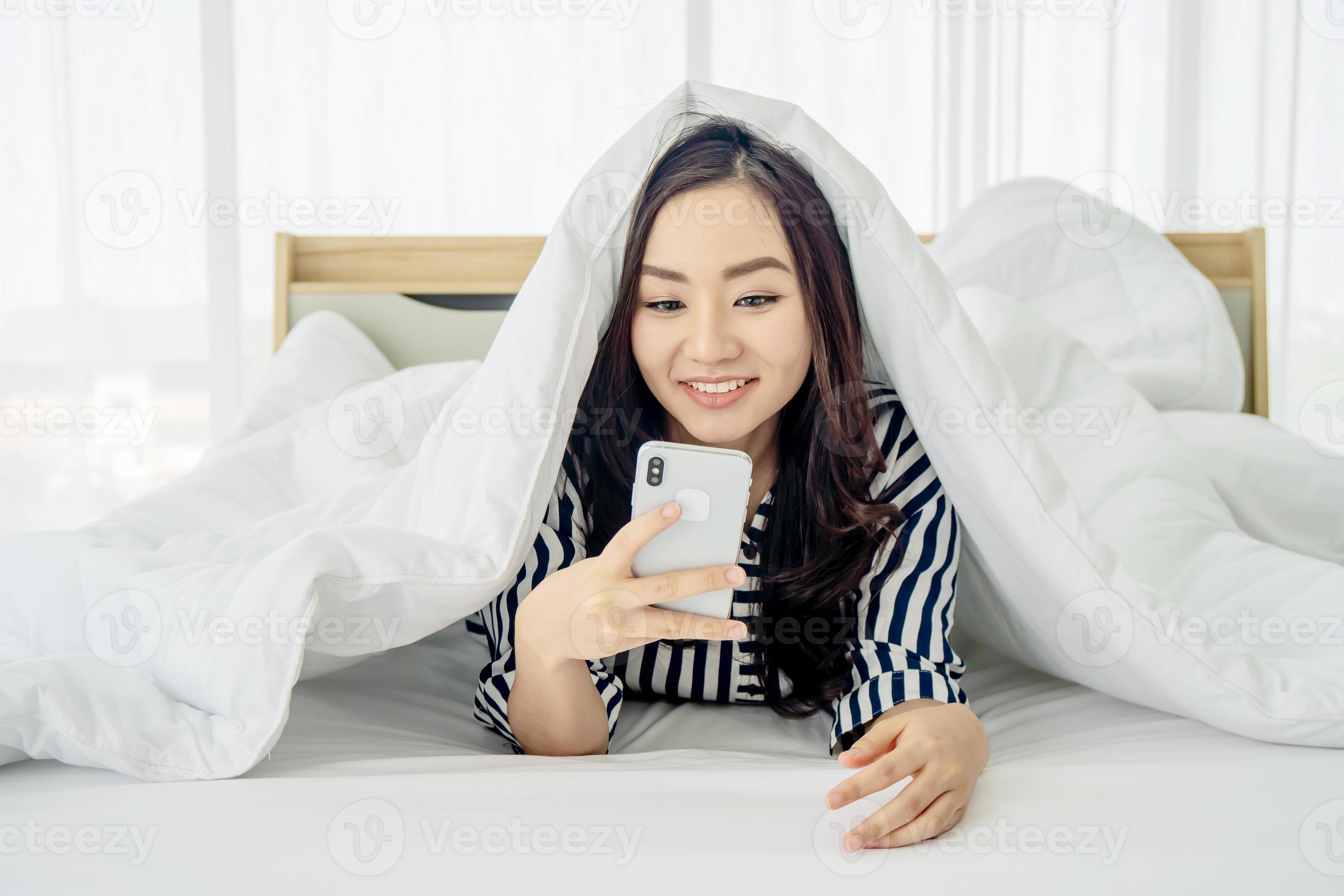 Photo of a happy pretty young woman using mobile phone under blanket in