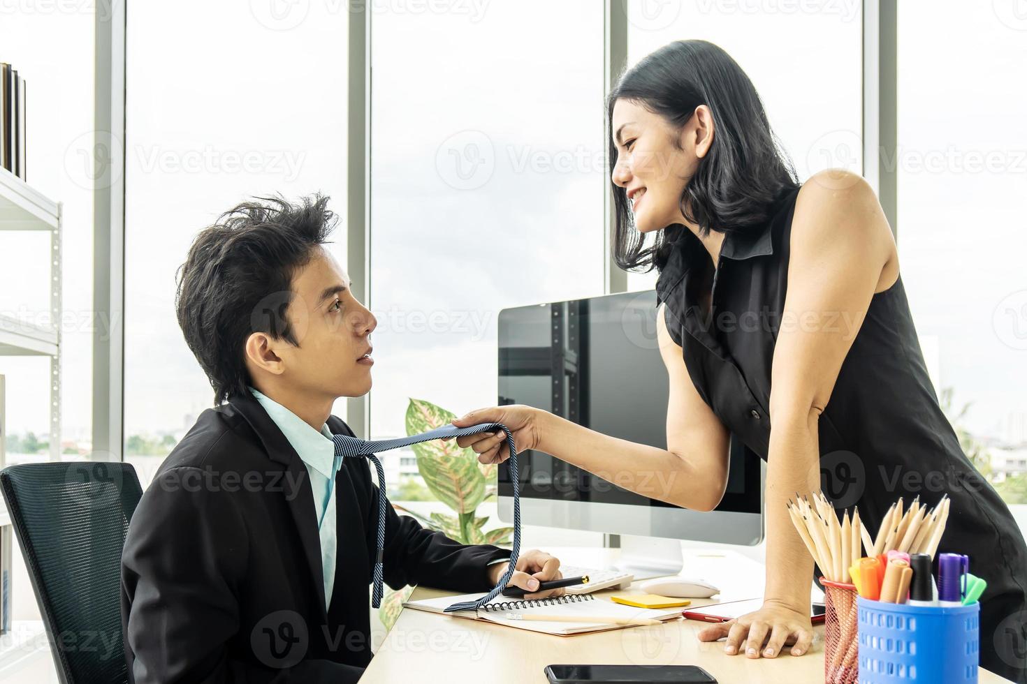 seduciendo a colegas en la oficina. mujer atractiva y elegante con un vestido negro parada y sosteniendo su corbata frente a un hombre con traje, mirándolo a los ojos foto