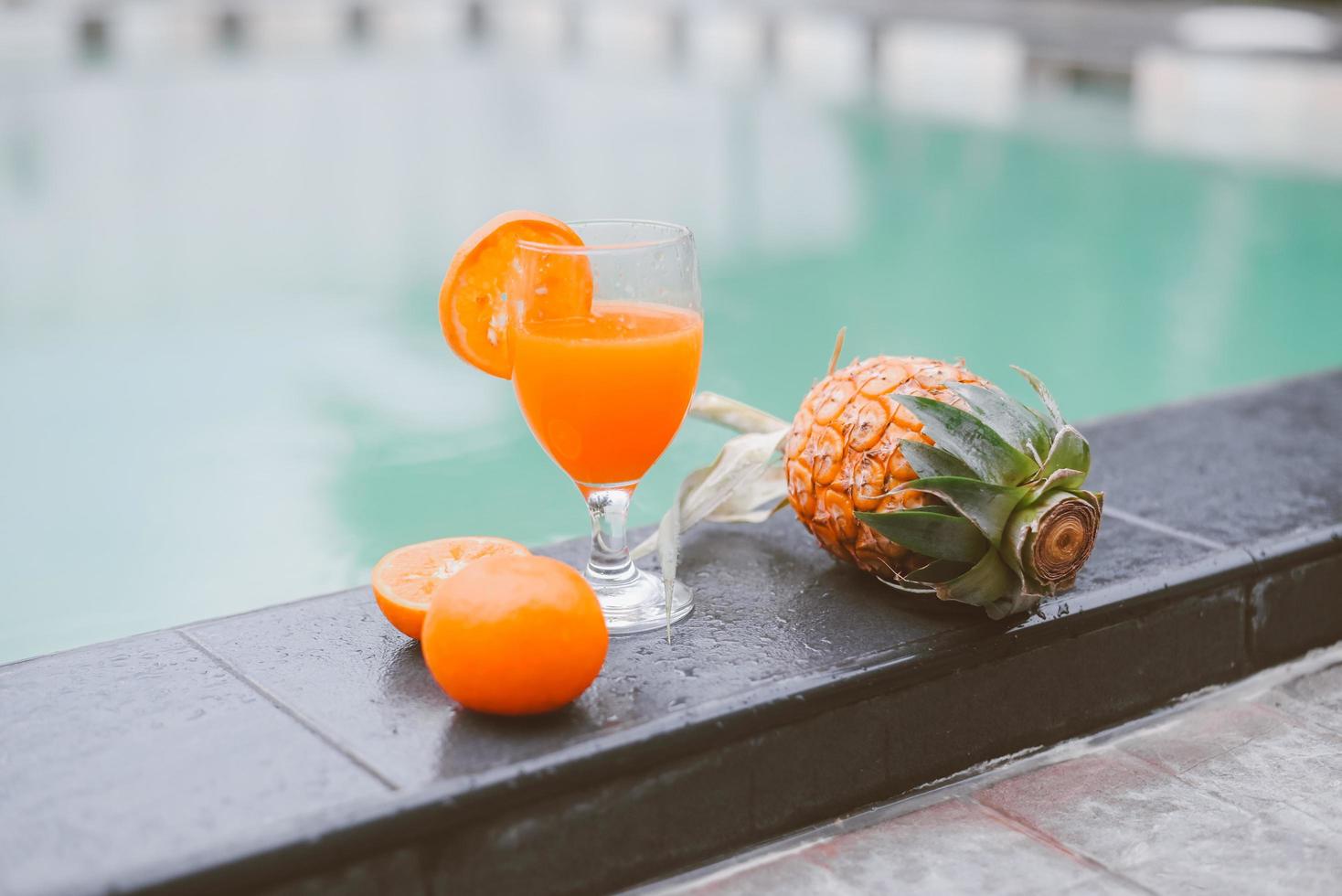Glass of juice on pool edge, orange juice with orange fruit and pineapple on the swimming pool photo