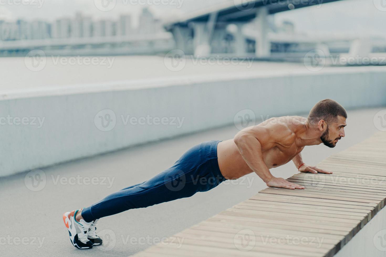 Horizontal shot of muscular athlete man does push up exercise has strong body stands in plank pose outdoor wears sport trousers and sneakers concentrated into distance. Strength and motivation concept photo