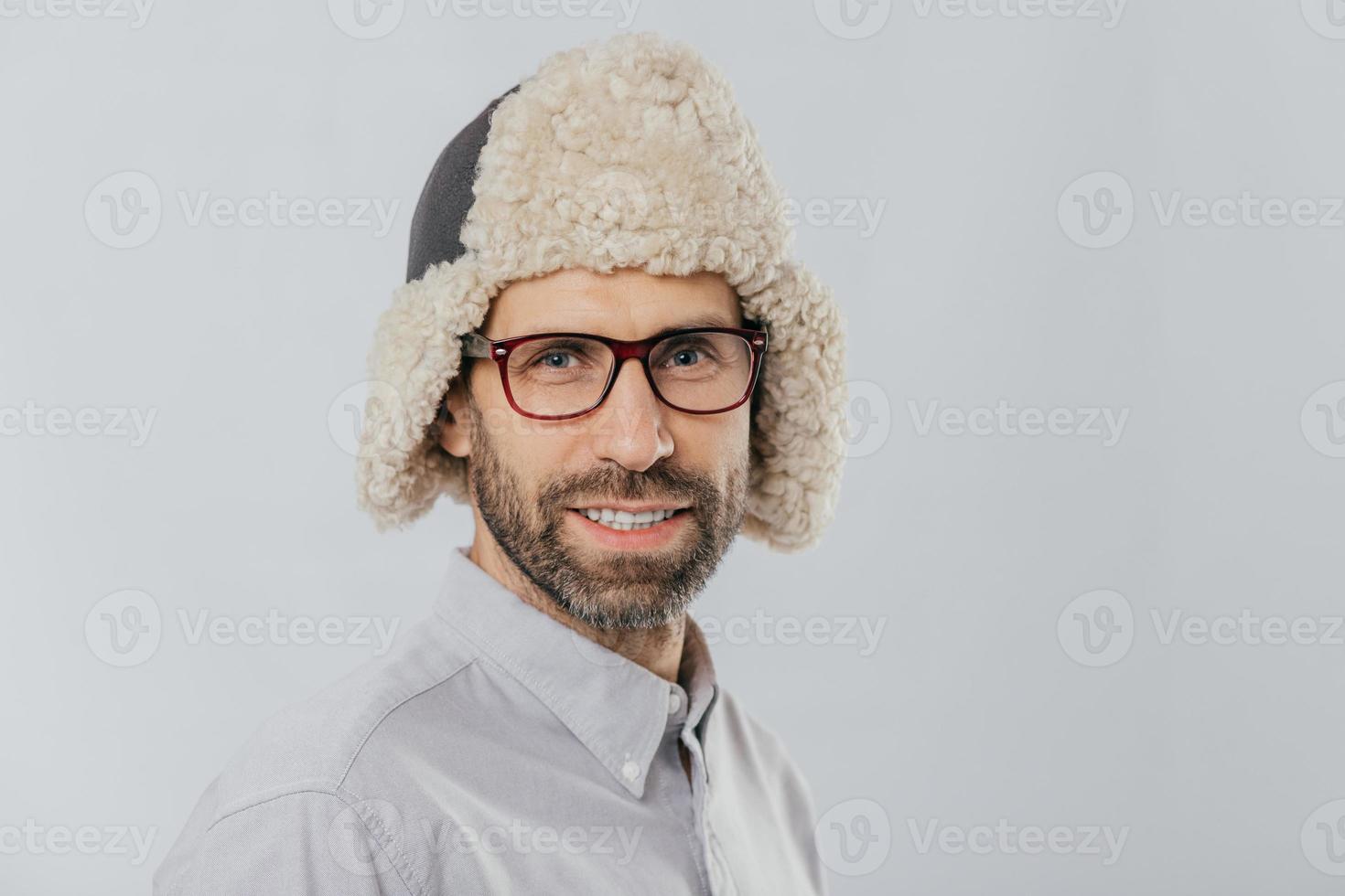 Photo of attractive young European male with pleasant appearance, wears warm fut hat, transparent glasses, models over white studio wall, rejoices shopping during weekend. People, style concept