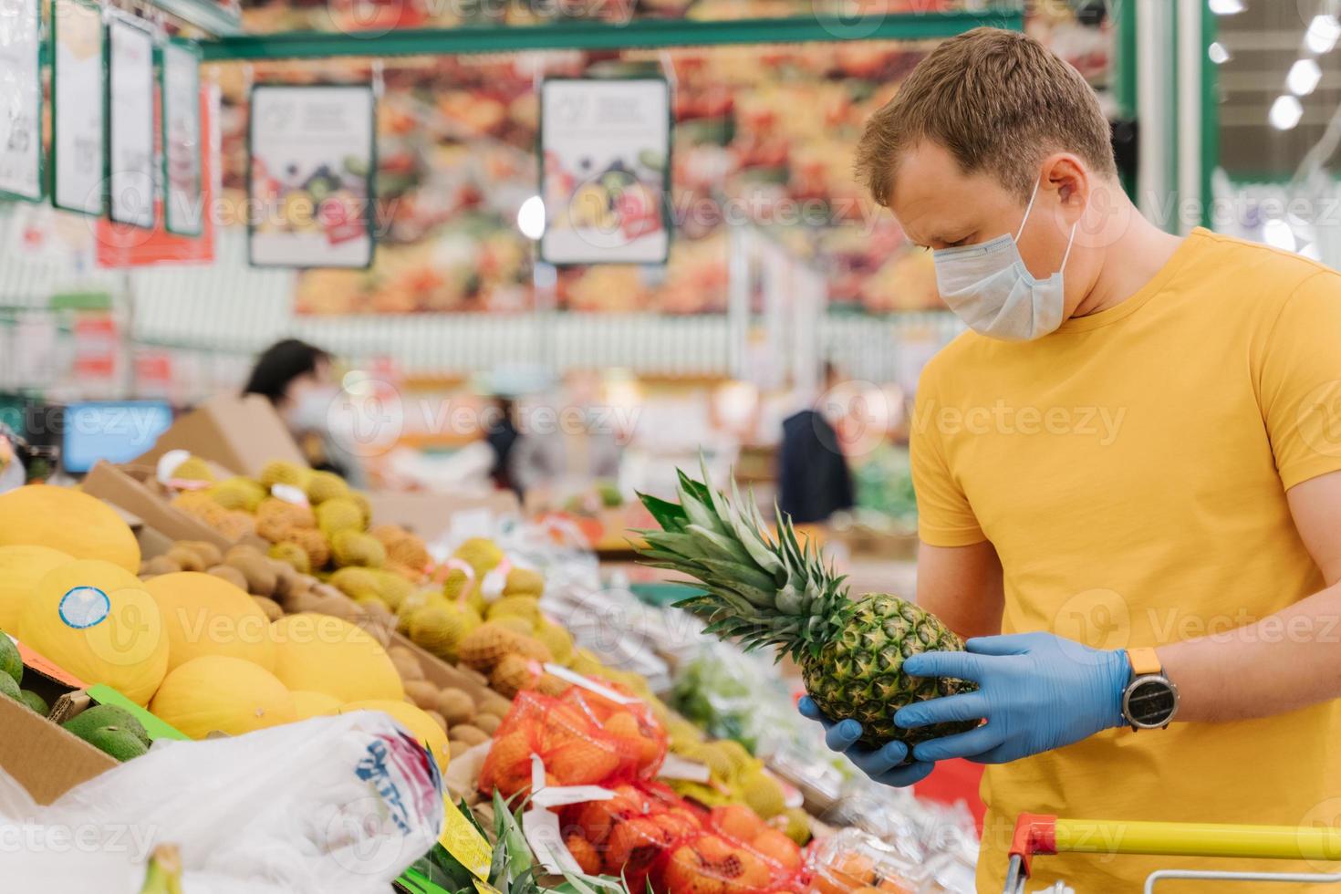 Horizontal shot of man selects pineapple in store wears medical mask and gloves buys fresh fruit during coronavirus outbreak or virus pandemic poses in grocery department. Protection, prevent measures photo