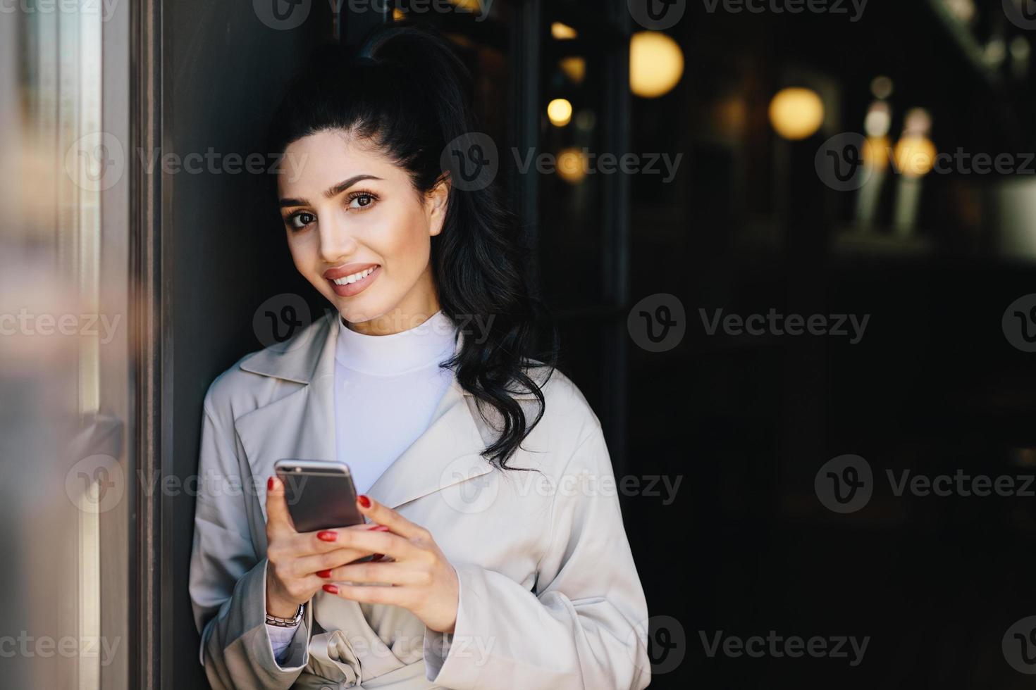 mujer morena sonriente con cola de caballo con manicura roja vestida formalmente sosteniendo un teléfono inteligente haciendo compras en línea mientras espera el tren. mujer de negocios con teléfono celular yendo a trabajar foto