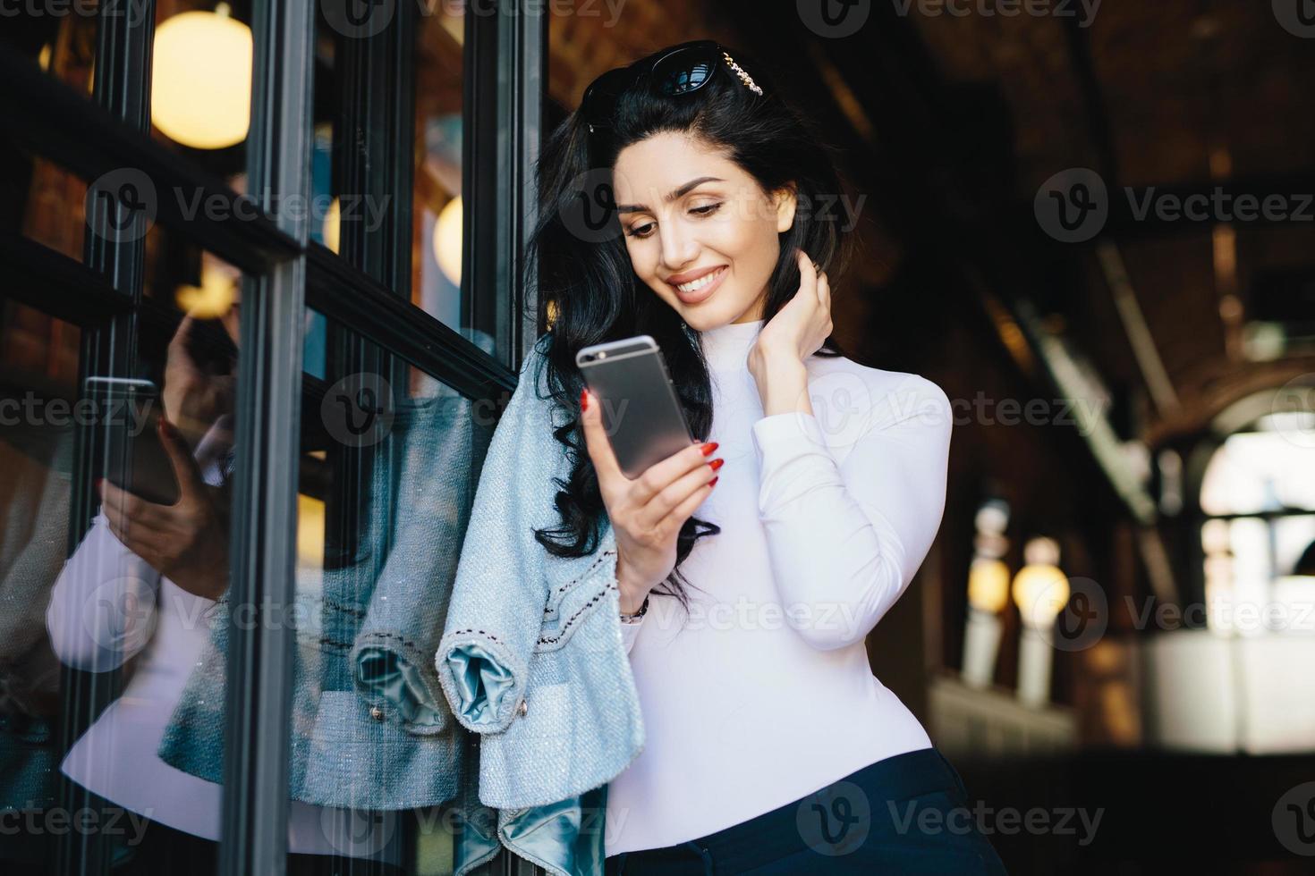 joven empresaria morena pequeña con ropa elegante parada en el interior del café esperando que la gente se reúna usando teléfonos inteligentes en el interior, enviando mensajes de texto en línea. concepto de comunicación foto