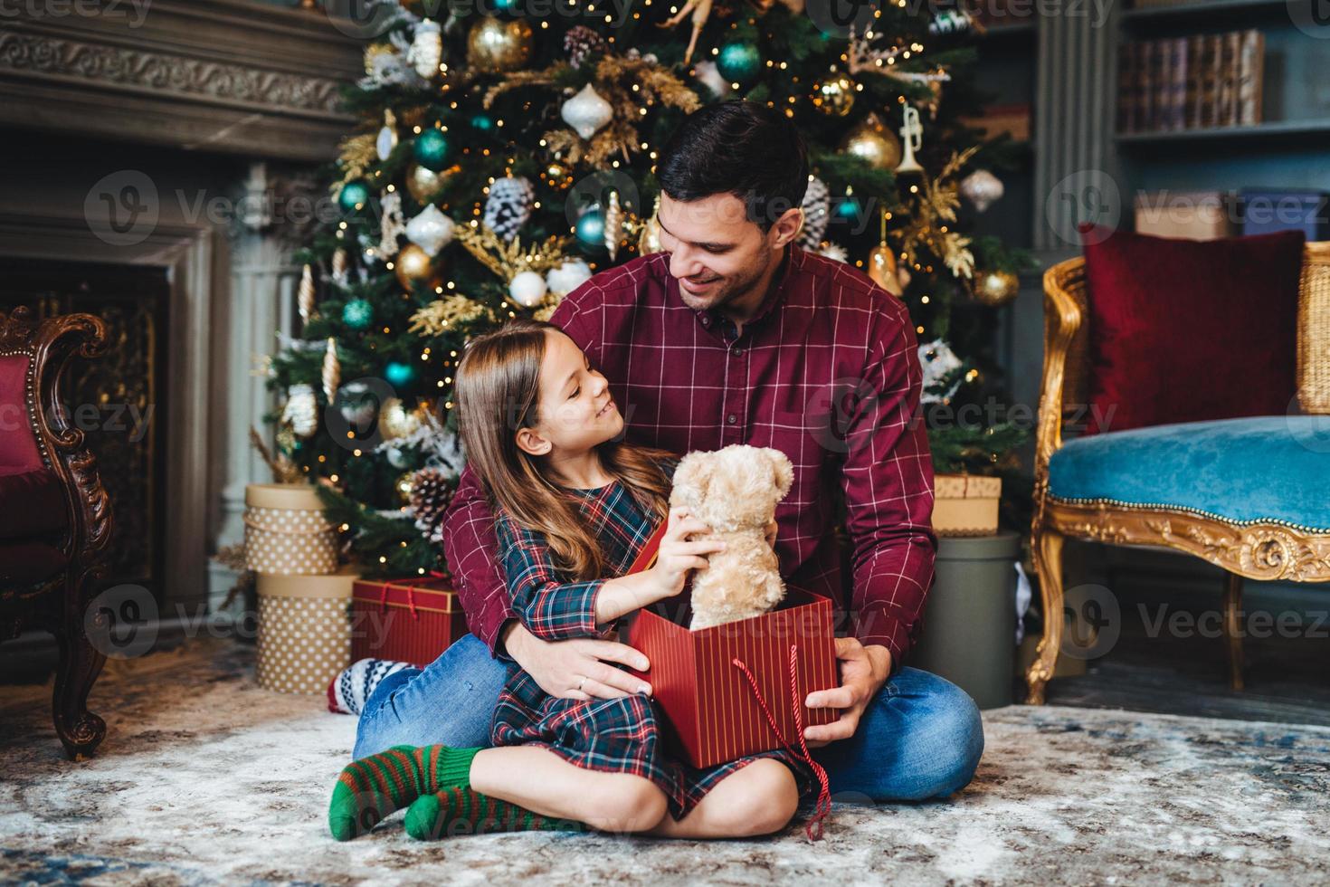 Small female child holds teddy bear, recieves unexpected gift from affectionate father, being thankful to him. Young dad gives present to daughter, congratulates her with Christmas or New Year photo