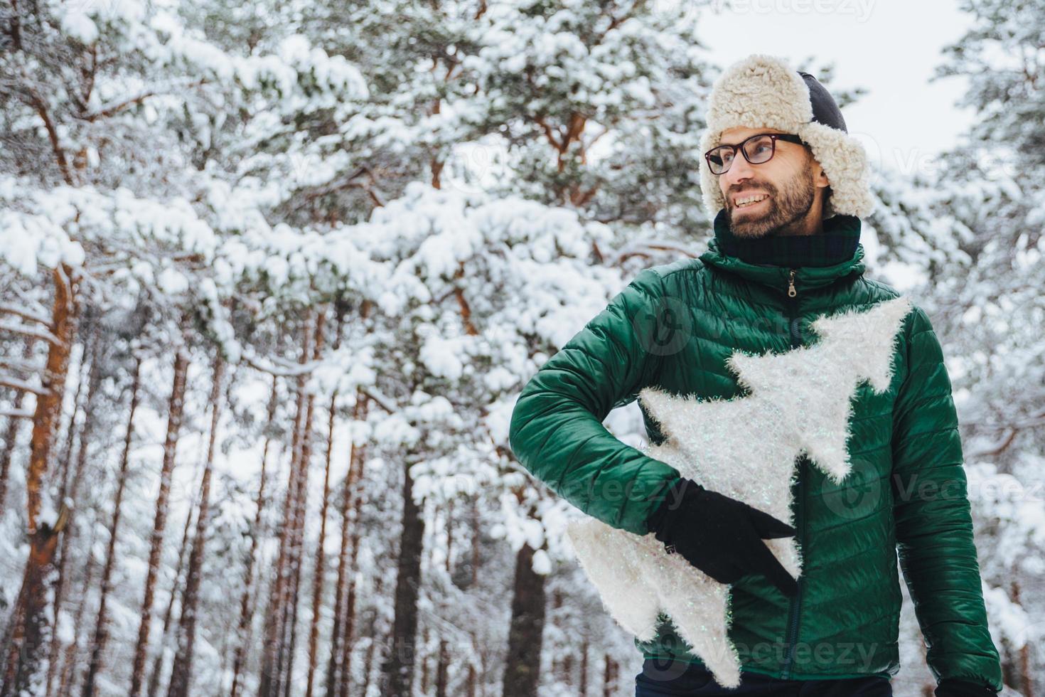 un hombre alegre y agradable con barba mira cuidadosamente a un lado, sostiene un abeto, usa gafas, sombrero y chaqueta, posa contra los árboles de invierno cubiertos de nieve. concepto de personas y estilo de vida foto
