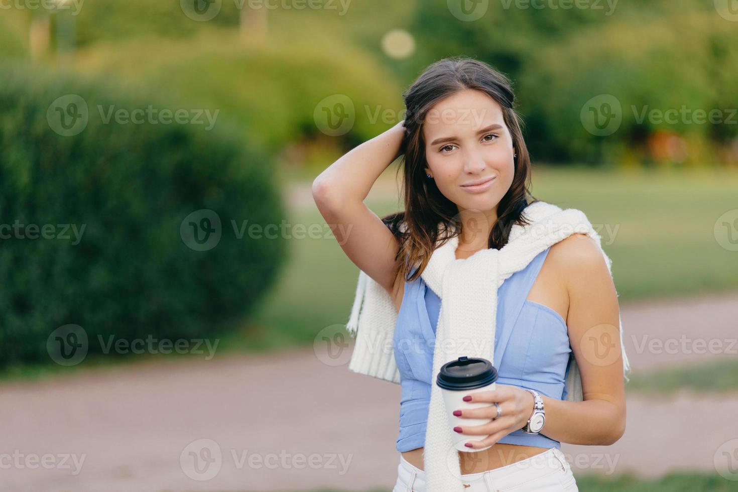 Pleased beautiful European woman with fit figure, keeps hand on head, drinks takeaway coffee, stands against green blurred background with copy space for your advertisement. Coffee time concept photo