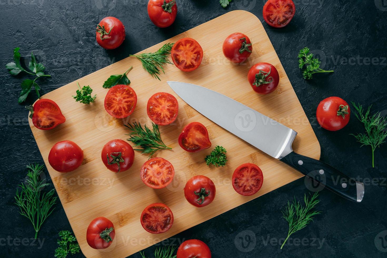 Top view of fresh small red tomatoes sliced on wooden kitchen board with knife. Green parsley and dill near. Vegetables and vitamins photo