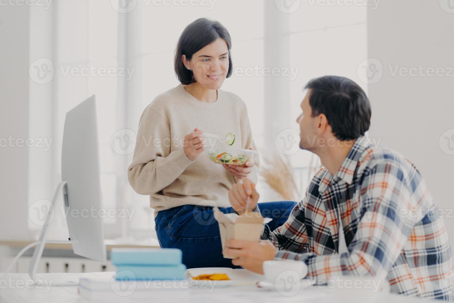 Photo of lovely woman and man discuss working issues together while have lunch, eat fast food and fresh vegetable salad, pose at desktop with computer monitor and scientific literature. Teamwork