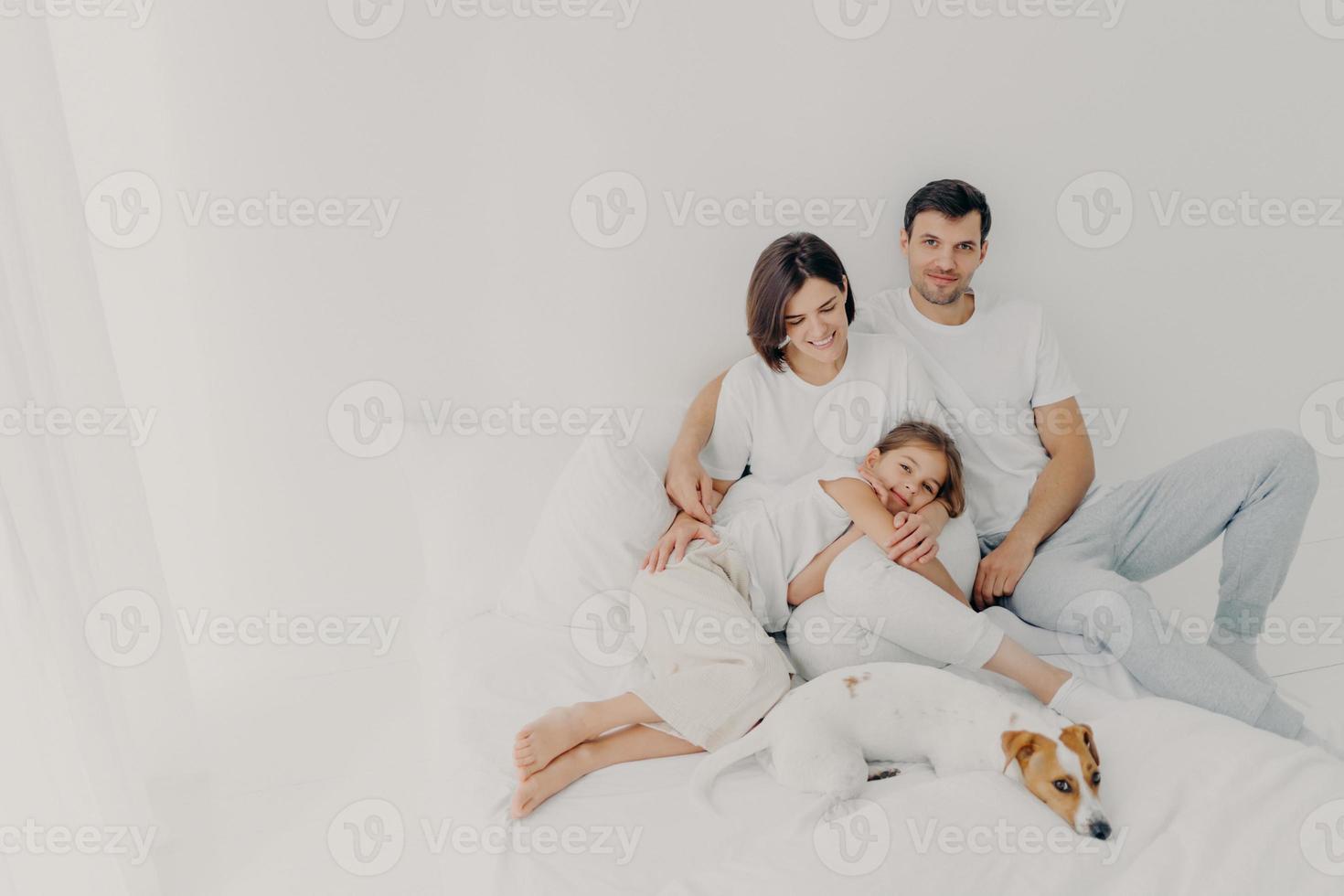 feliz pose familiar en la cama blanca durante el fin de semana. el padre, la madre, su hija y el perro se sienten cómodos al estar en el dormitorio, abrazarse y divertirse, estar relajados y alegres. familia amistosa y descanso foto