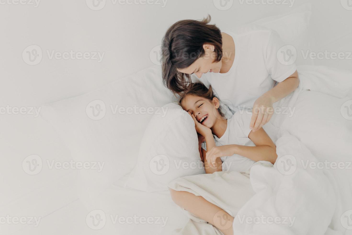 Overjoyed mum and daughter have fun together in bedroom, pose under soft blanket. Happy small girl spends leisure time with mother, has toothy smile, expresses happiness. Children and parents photo