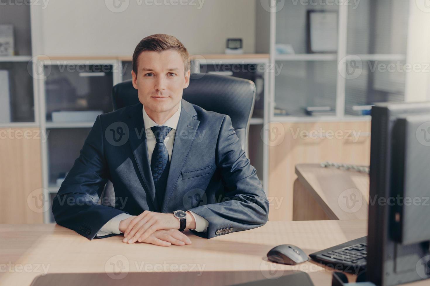Horizontal shot of prosperous young businessman in elegant suit, sits at work place, surrounded with computer, has confident look directly into camera, being professional in business sphere. photo
