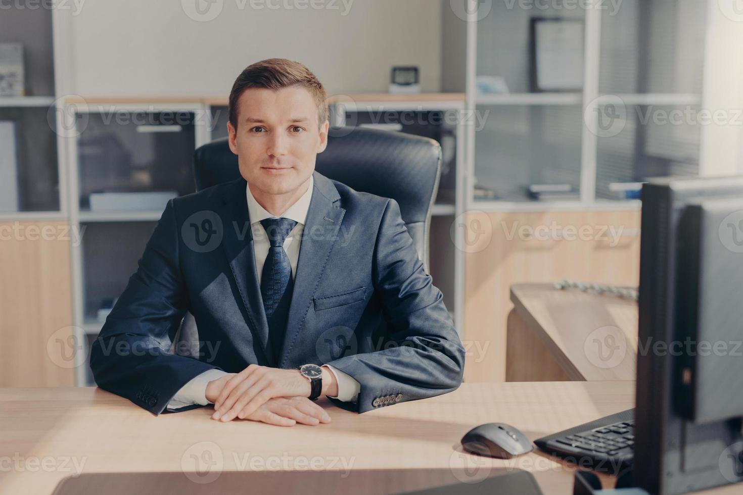 toma horizontal de un próspero joven hombre de negocios con traje elegante, se sienta en el lugar de trabajo, rodeado de una computadora, tiene una mirada segura directamente a la cámara, siendo profesional en el ámbito empresarial. foto