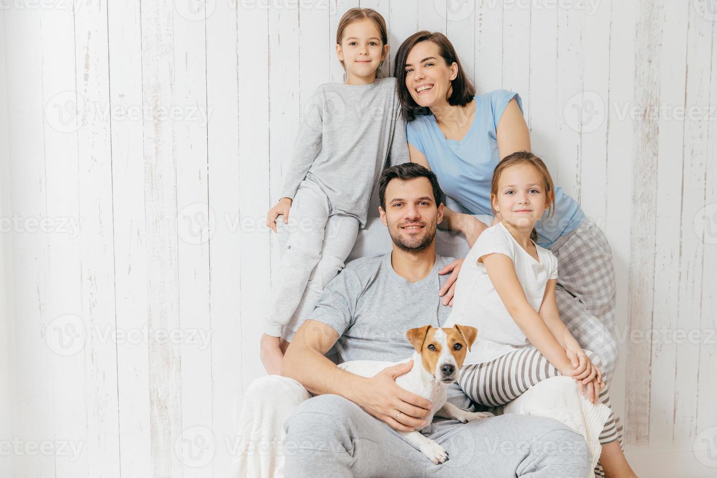 Horizontal shot of friendly family pose together against white background two little sisters, father, mother and their pet. Happy parents and their female children. Family of four. Parenthood photo