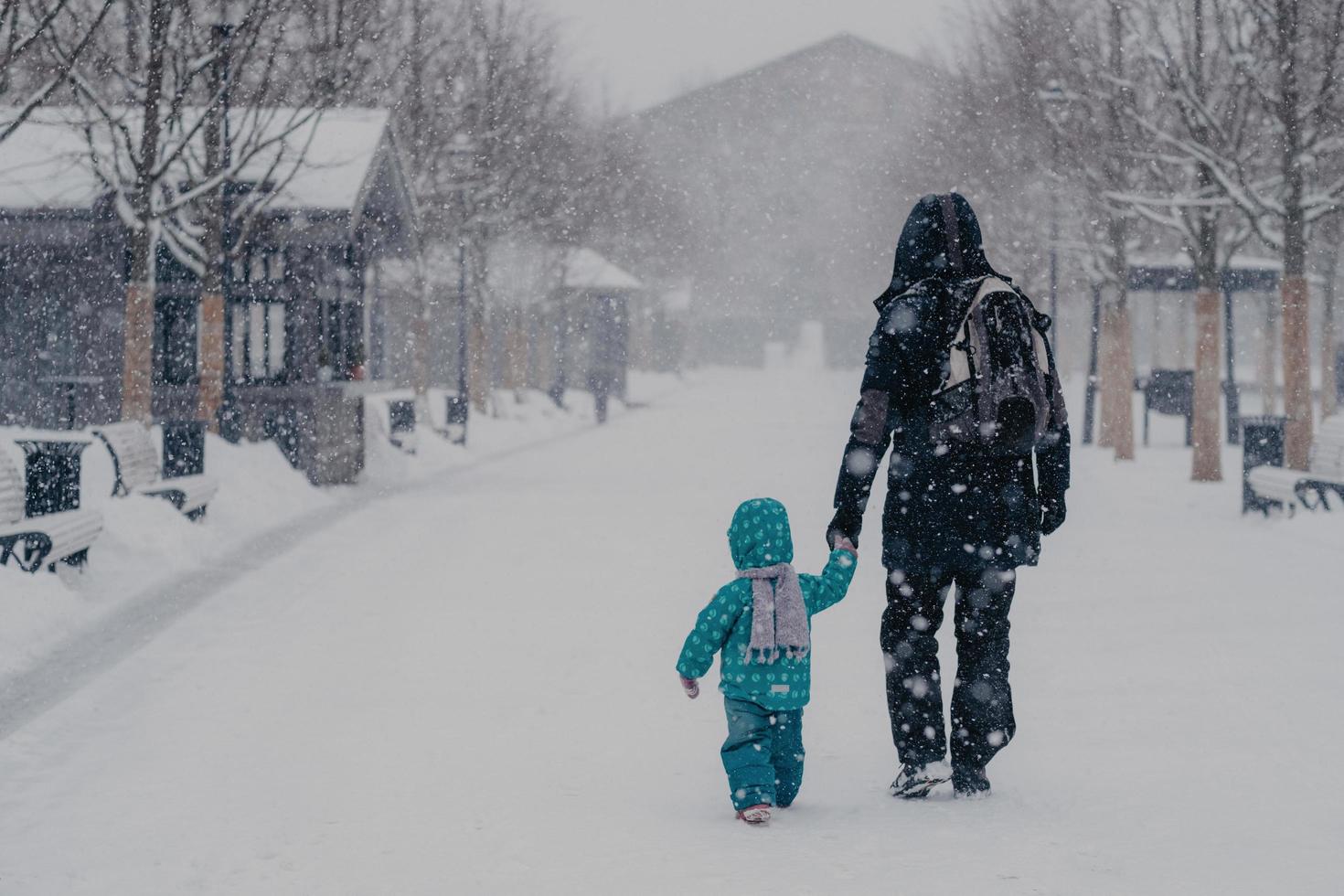 Back view of affectionate father and little male child hold hands, walk along snowy street during heavy snowfall in winter, enjoy recreation time, go home, dressed in warm clothes, carry rucksack photo