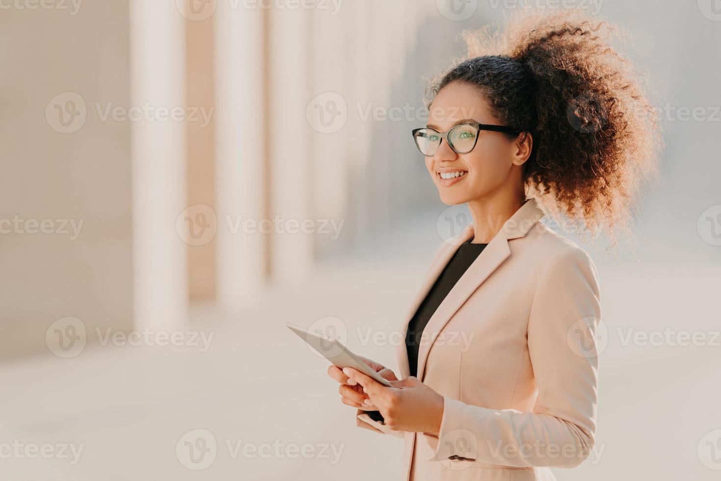 Profile shot of glad dark skinned woman with Afro hair, dressed elegantly holds tablet computer in hands strolls at street wears optical glasses connected to high speed internet. Lifestyle, technology photo