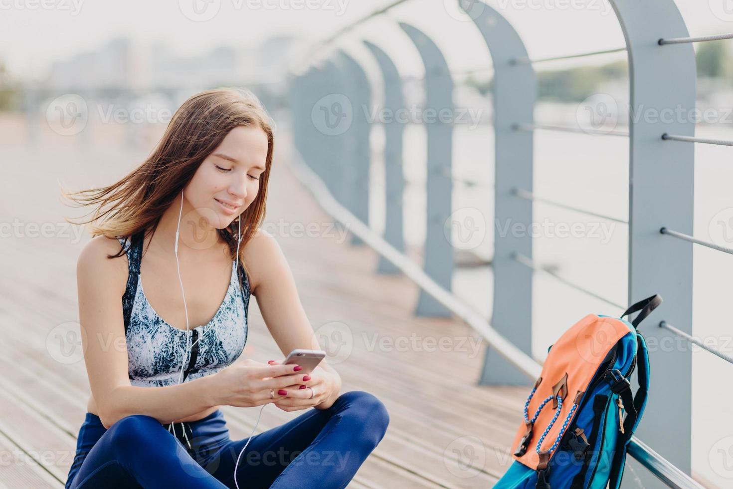Positive fitness girl in top and leggings, has glad expression as reads pleasant text message recieved from boyfriend, has dark hair, rests outdoor. People, sport, modern technologies and lifestyle photo