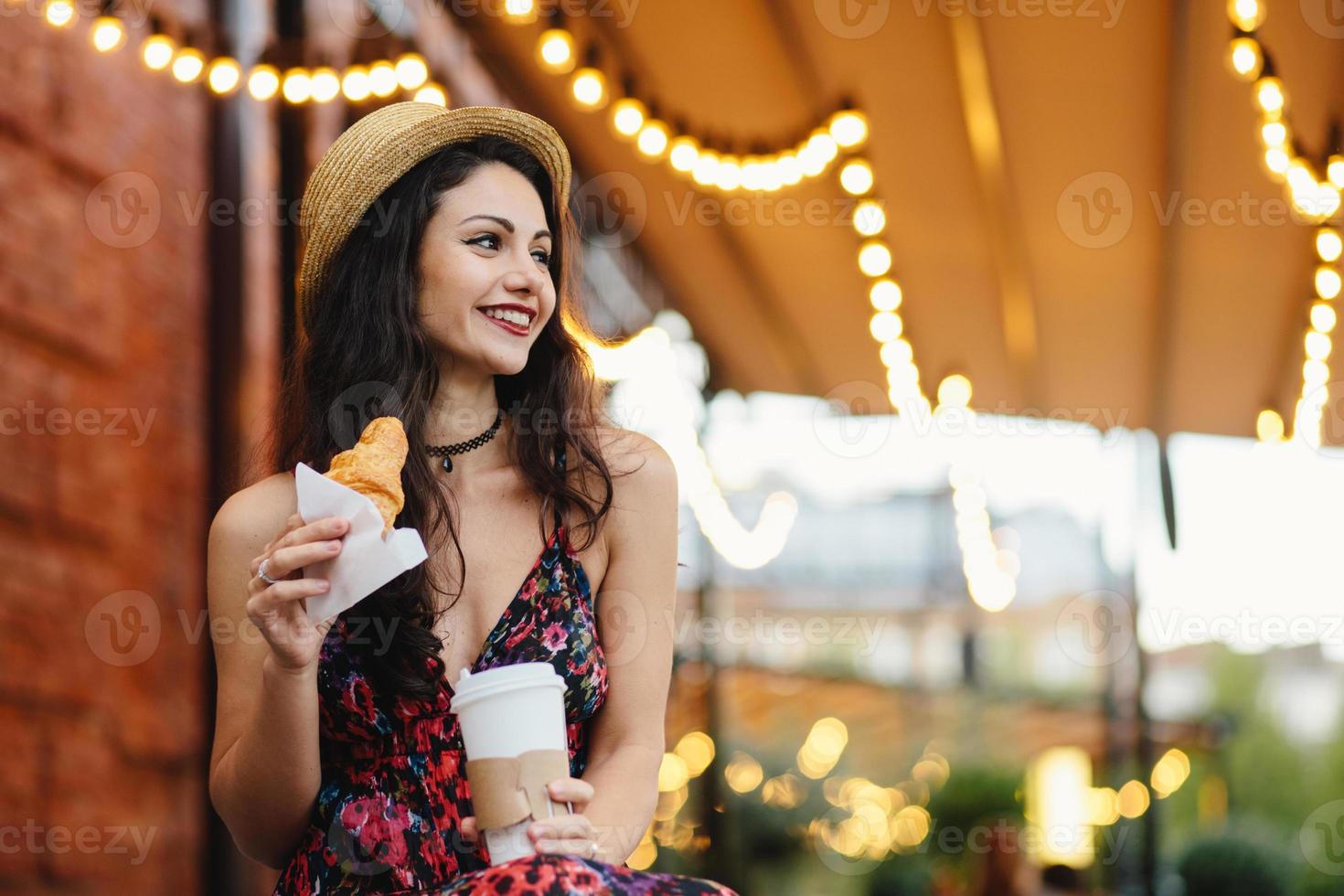 Delightful brunette female with pleasant appearance wearing summer hat and dress holding croissant and takeaway coffee, resting at terrace looking aside with happy expression. People, recreation photo
