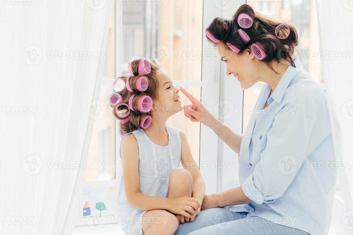 Horizontal shot of pretty young mother and daughter with curlers on hair, being in good mood, enjoy time at home, prepare for holiday. Happy family concept. Beauty, children and fashion concept photo