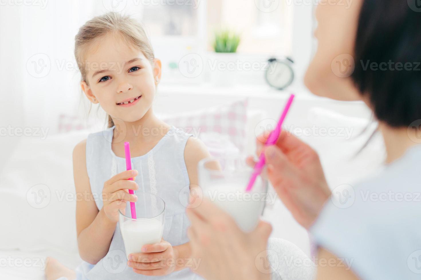 Pretty cute small child drinks delicious cocktail, has pleasant talk with her affectionate mother, enjoy together good morning, plan what to do during day off. Light tones. Spacious cozy bedroom photo