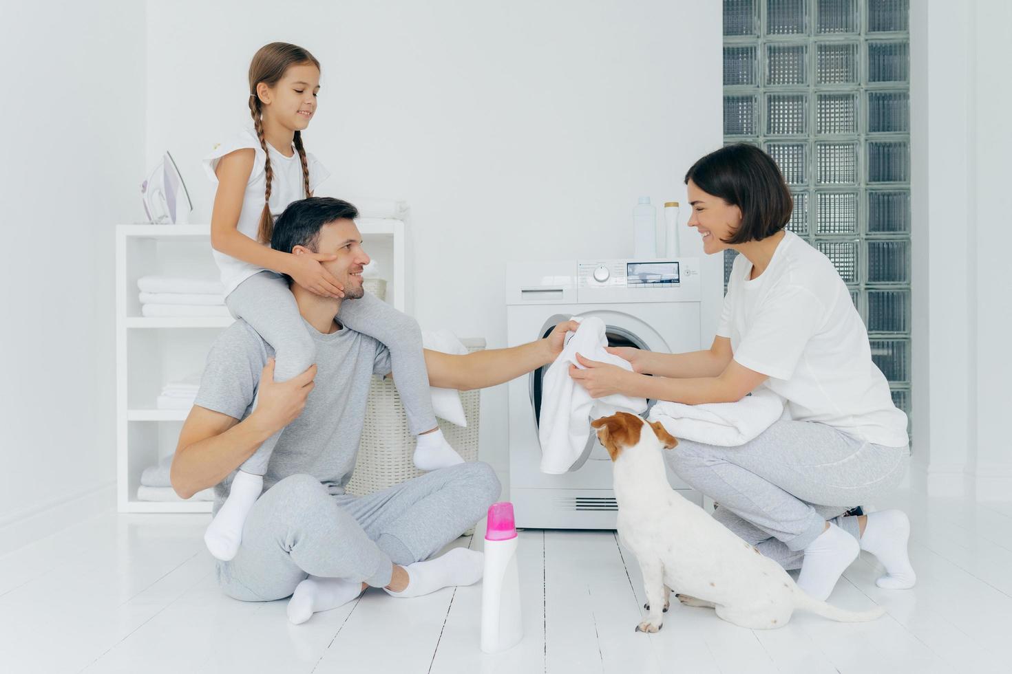 Happy father gives piggy back to daughter, helps wife to load washing machine, their pet poses near, bottle of washing powder in white floor. Friendly family in laundry room, wash linen, do housework photo