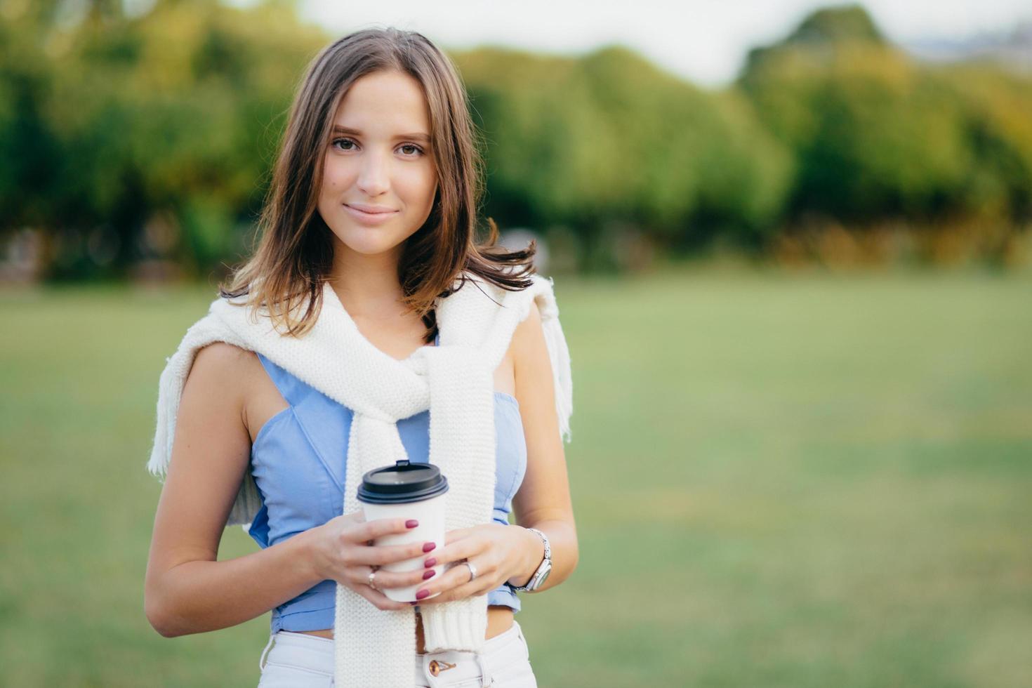 Photo of good looking woman with dark bobbed hairstyle, holds paper cup of coffee, has free time, spends weekend on nature, poses agaist green nature background with copy space for your text