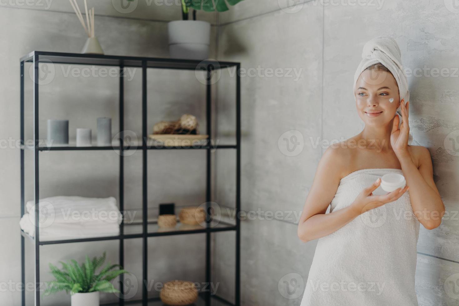 foto de una joven relajada que aplica crema facial, tiene una expresión satisfecha, envuelta en una toalla de baño, posa cerca de una pared gris en el baño, usa un producto cosmético para el rejuvenecimiento. belleza y cuidado de la salud