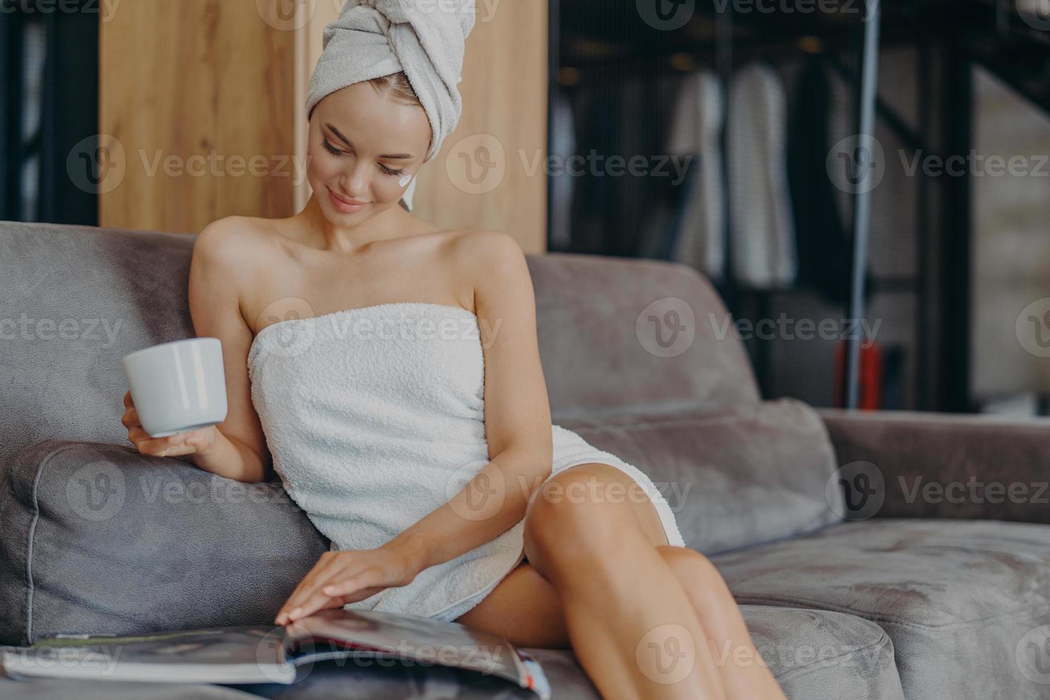 Indoor shot of relaxed beautiful woman sits relaxed on sofa, wears bath towel around body after taking shower, holds cup of tea, reads magazine, spends free time at home. Wellness, hygiene concept photo