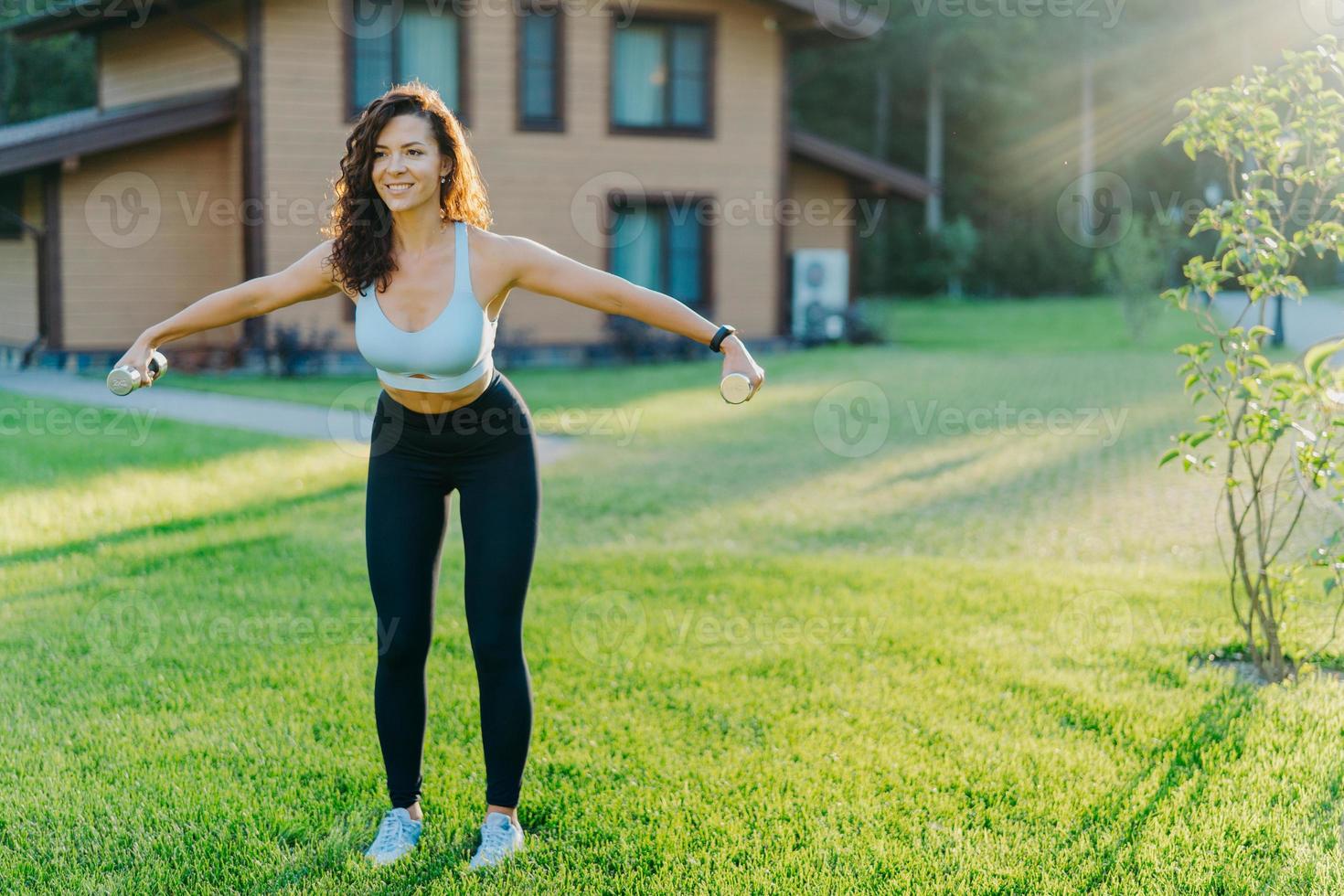 Slim motivated brunette woman dressed in cropped top and leggings, has workout with dumbbells, poses on green lawn near private house, has perfect body shape. Healthy lifestyle and sport concept photo