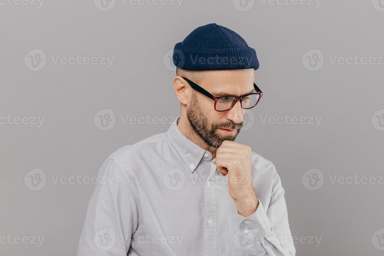 Indoor shot of concentrated man holds chin and focused down, being in deep in thoughts, thinks about innovative idea for his research, has clever expression, wears glasses and white formal shirt photo
