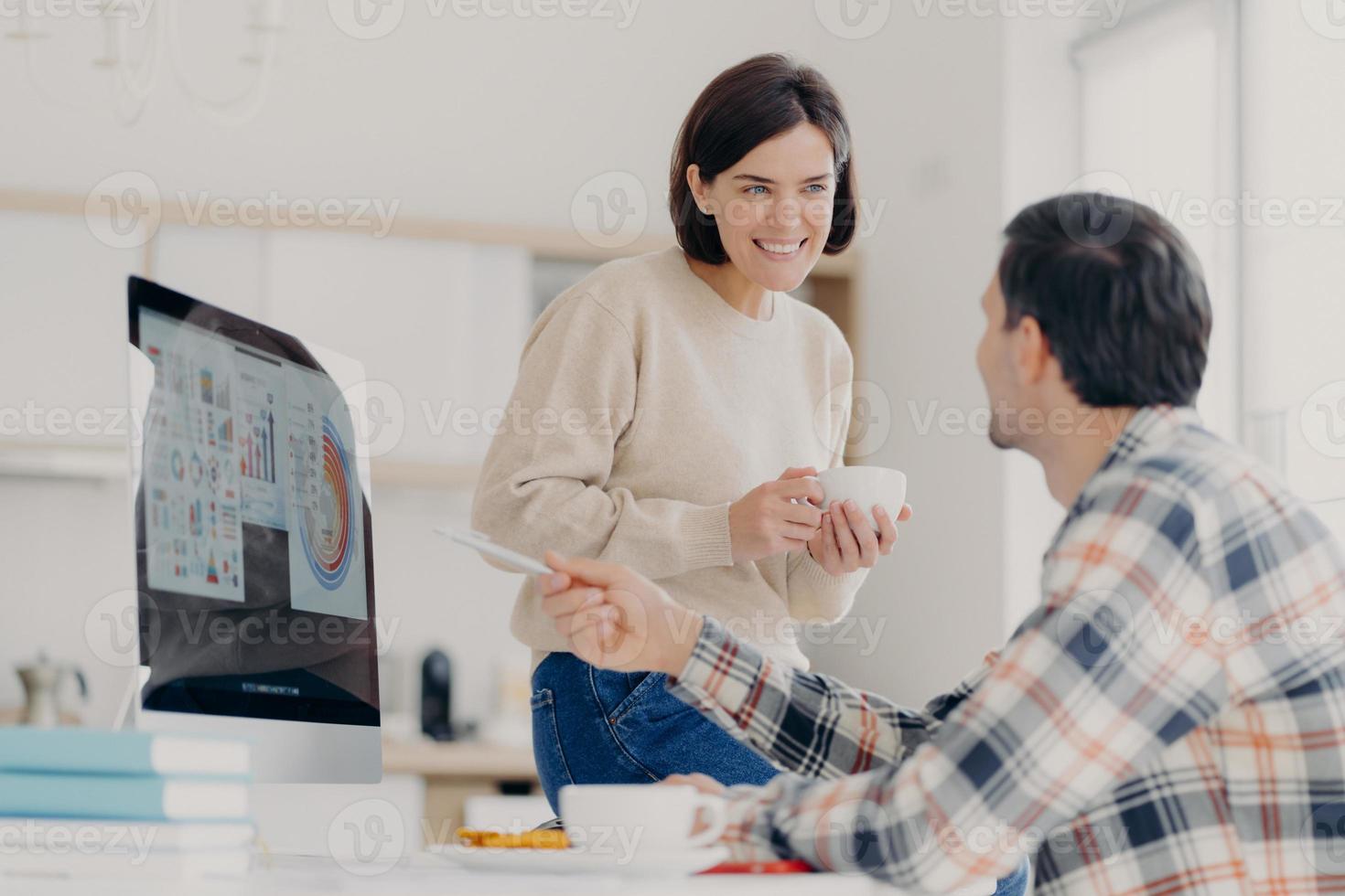 Positive woman and man discuss payment of utility bills during coffee break, man points into screen of computer, shows some graphics and diagrams, work at home in coworking space. Collaboration photo