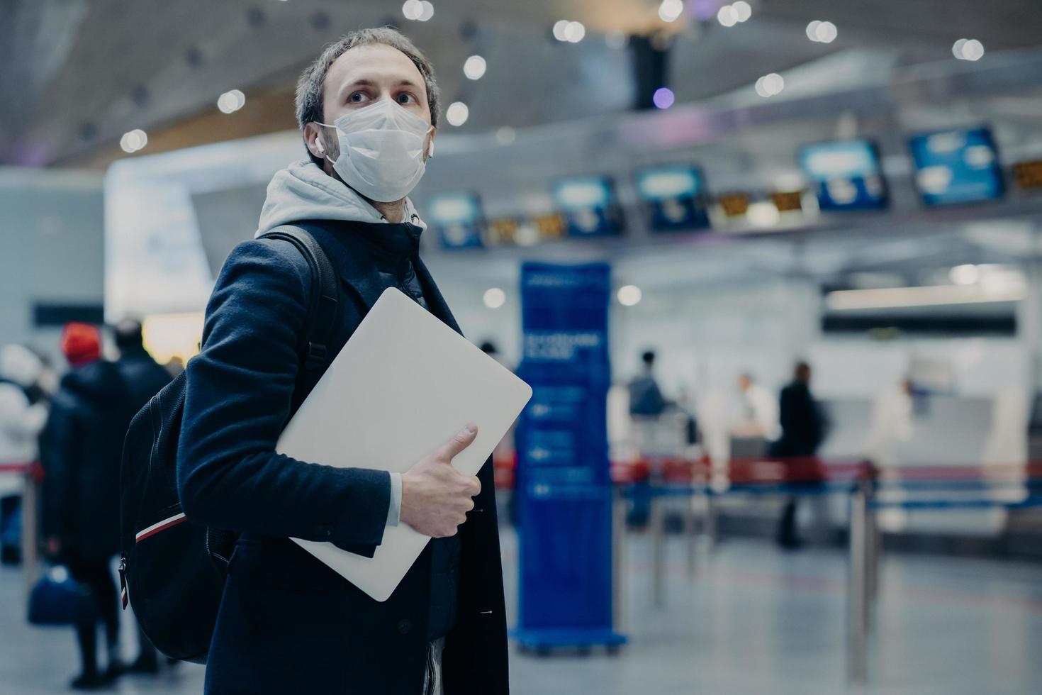 Man traveler wears protective disposable medical mask in airport, returns from abroad where coronavirus spreading, carries backpack, takes care of health, protects from virus, has postponed flight photo