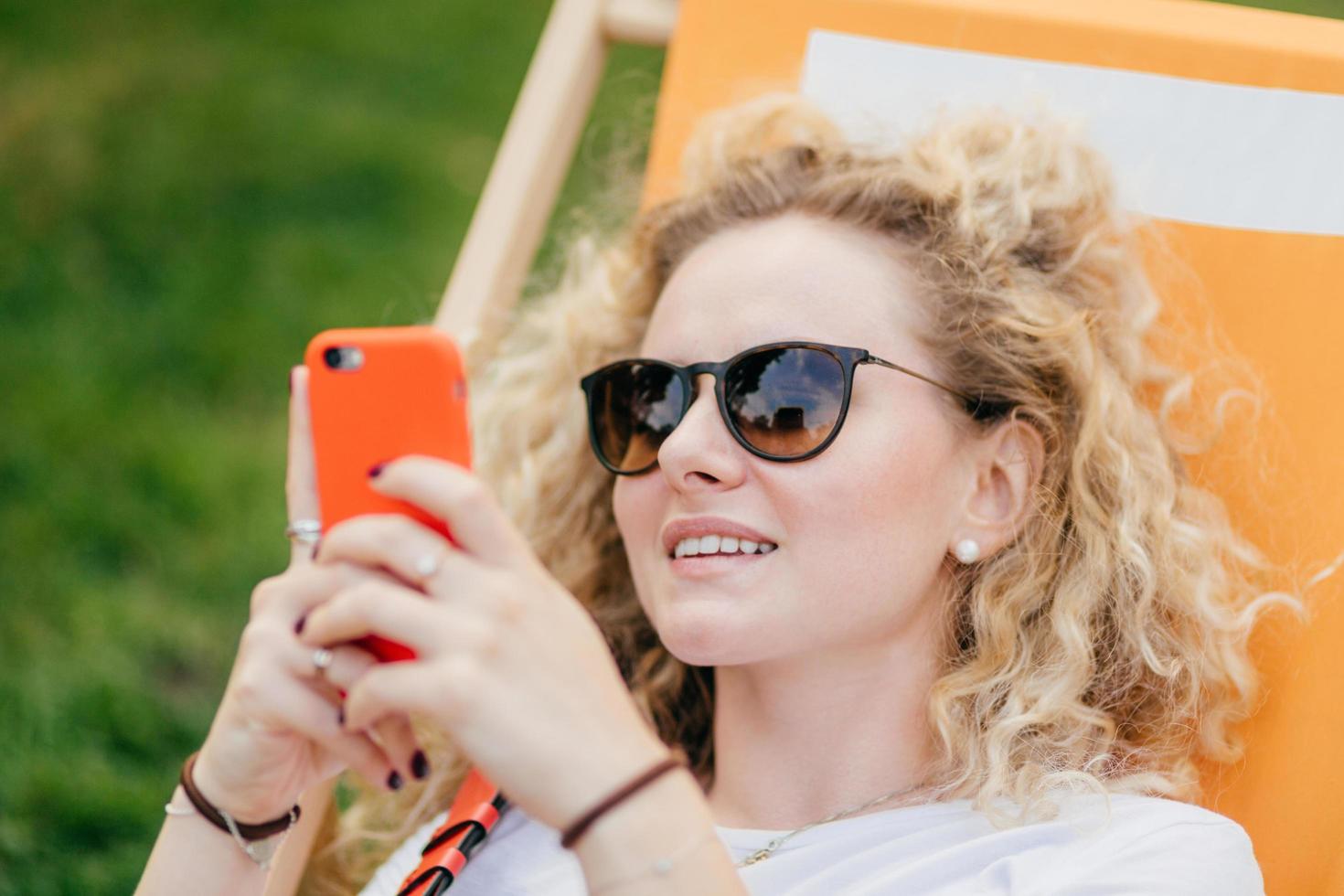 Outdoor shot of pleasant looking curly young female in sunglasses, holds modern orange smart phone, surfes social networks, connected to wireless internet, recreats in hammock during sunny day photo