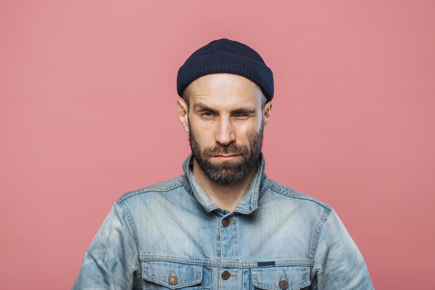 Photo of good looking bearded male blinks eye and looks seriously into camera, wears denim jacket and black hat, isolated over pink background. Stylish man with stubble poses in studio alone.