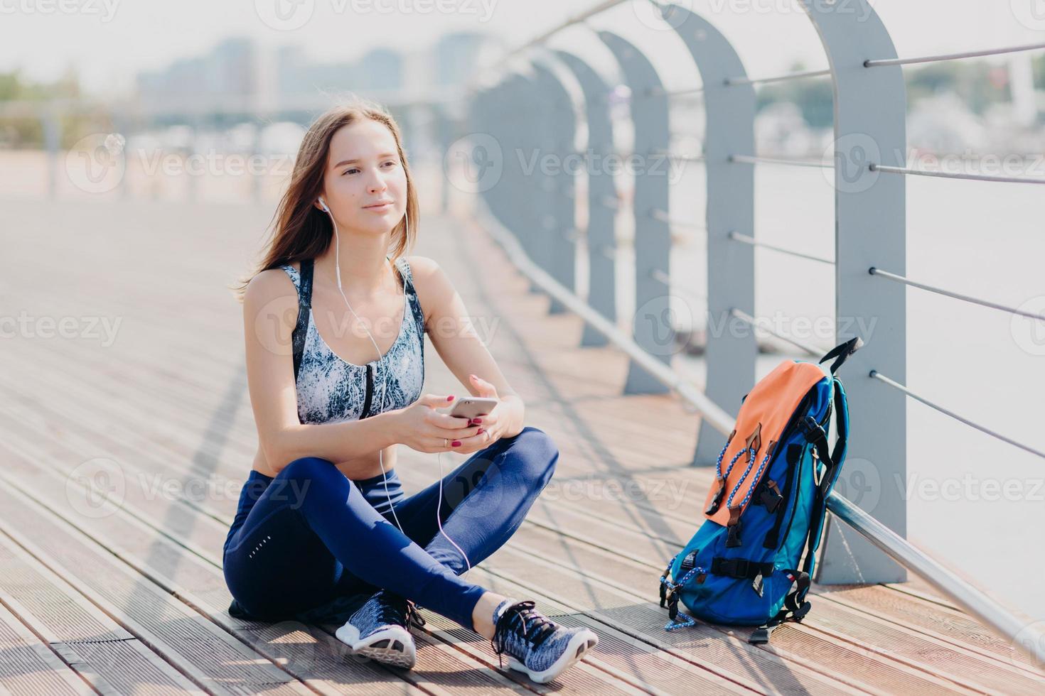 People and active lifestyle concept. Attractive young female enjoys calm atmosphere in urban territory, breathes fresh air, sits crossed legs, listens favourite music from playlist, uses mobile phone photo
