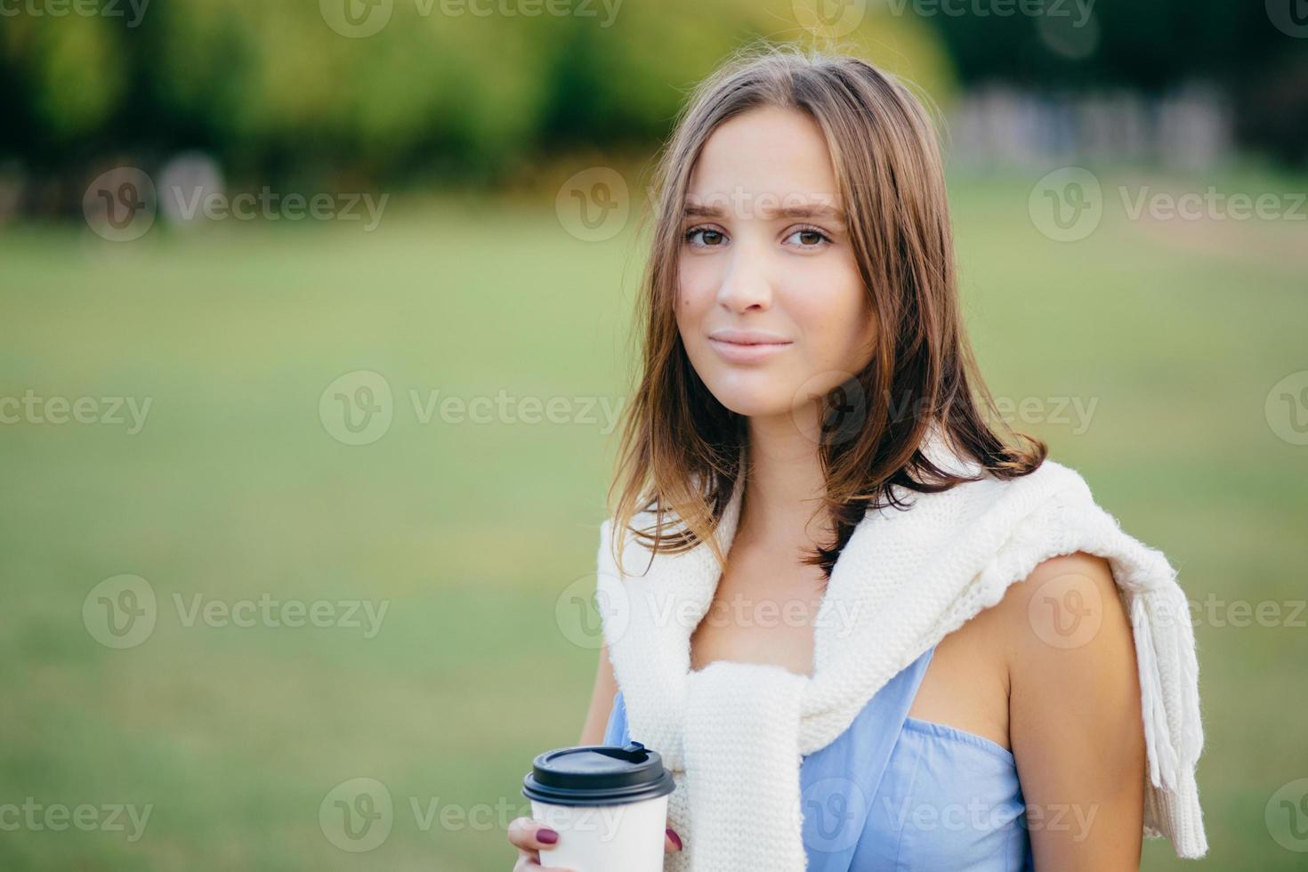 Photo of beautiful comely girlfriend with white sweater on shoulders, drinks takeaway coffee, thinks about something, stands outdoor, has walk across green field. People, lifestyle and spare time