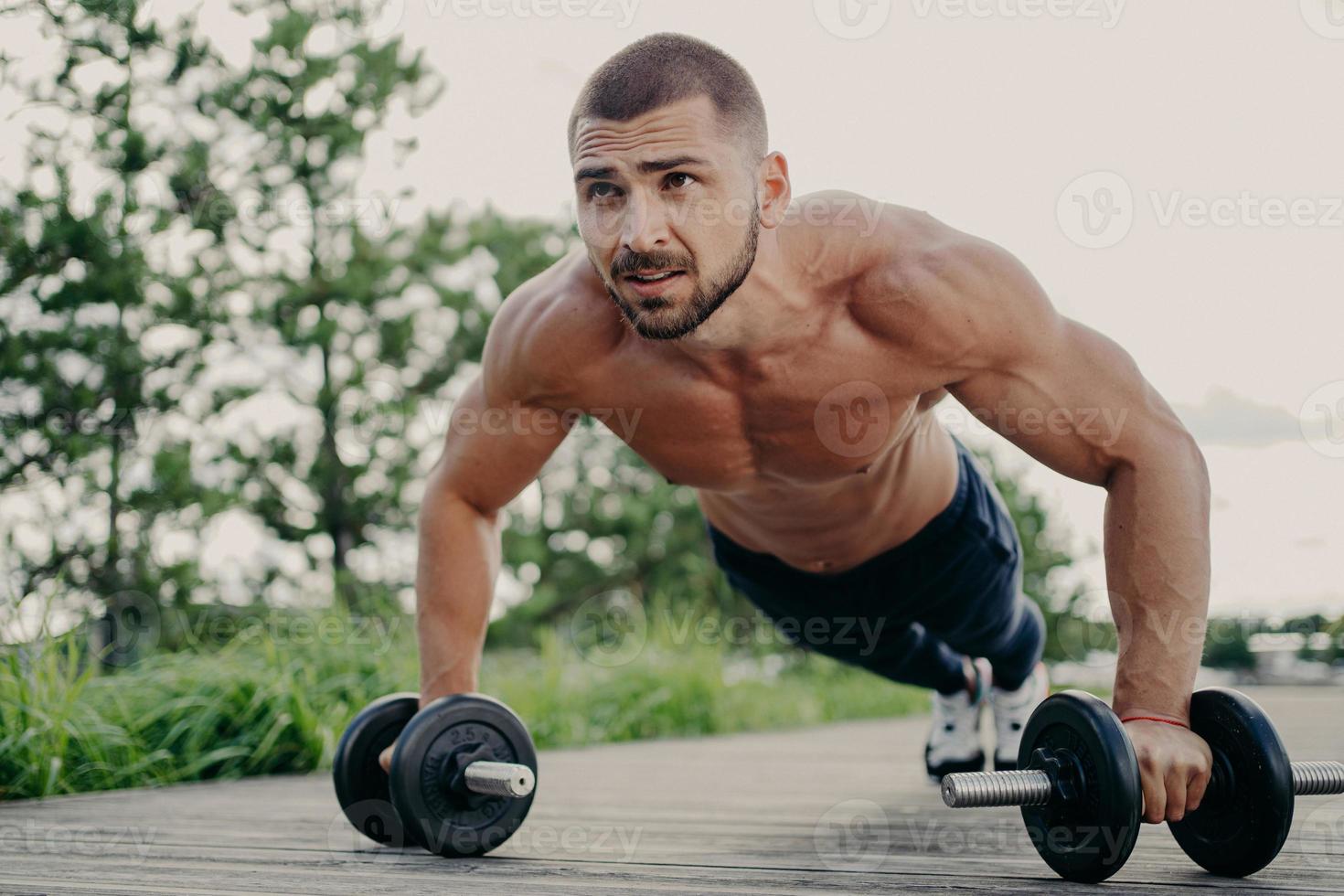 Young handsome bearded sportsman works out outdoor poses in plank with barbells, has strong body, muscular arms. Bodybuilder does exercises in open air, performs weight lifting, push up exercises photo