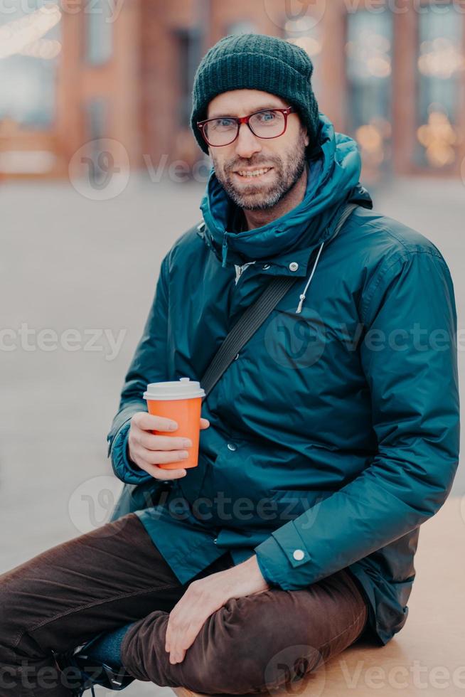 Stylish Caucasian man has free time, poses outside with disposable cup of coffee, dressed in hat and jacket, enjoys good rest, waits for someone at street, has satisfied expression. Lifestyle concept photo