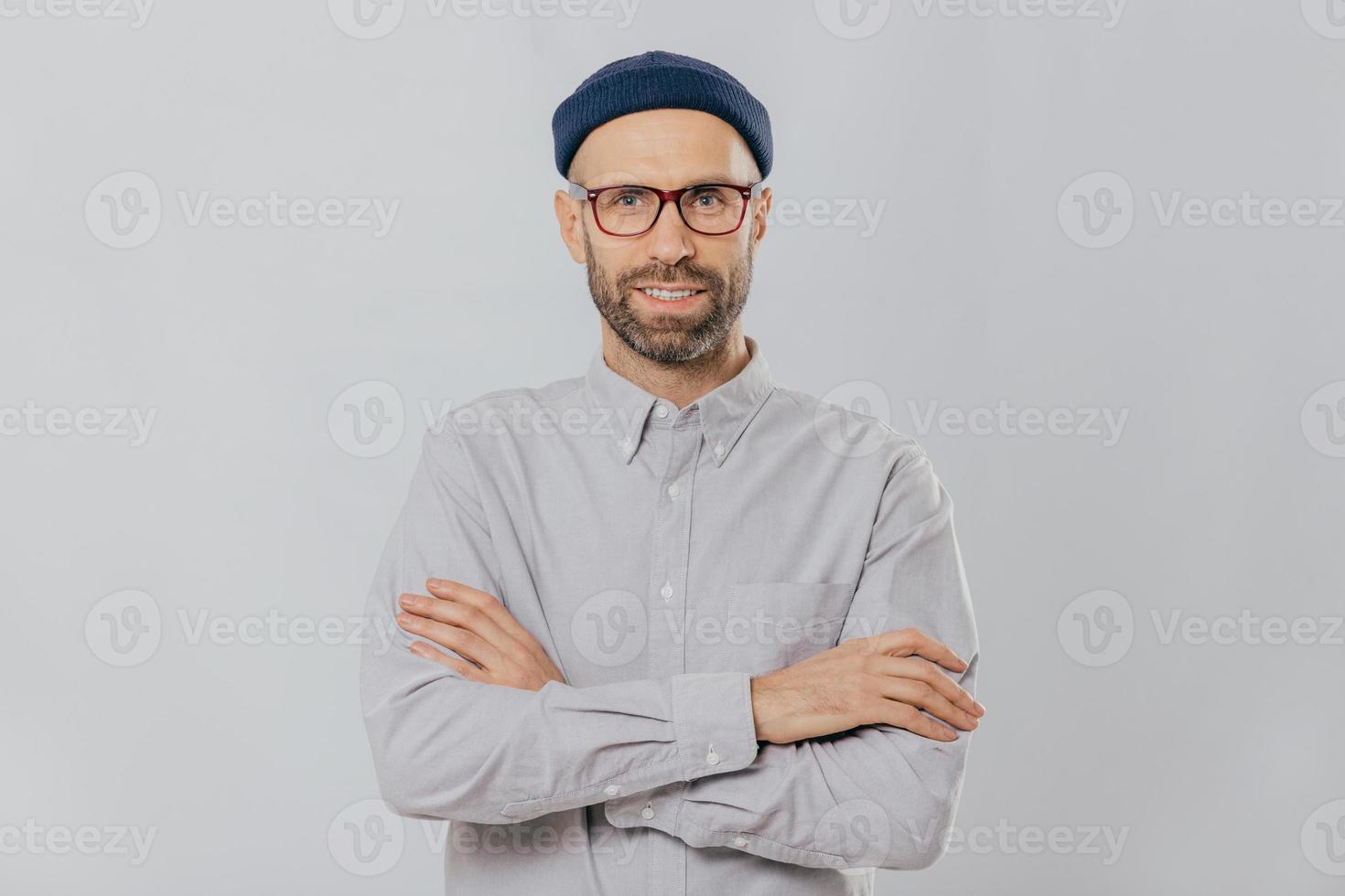 Horizontal shot of satisfied self confident male designer wears stylish headgear, dressed in white shirt, keeps arms folded, has dark stubble, isolated over white background. People and style concept photo