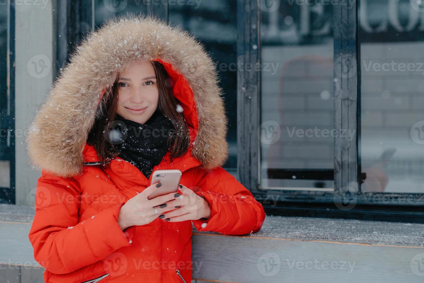 mujer joven de aspecto agradable con chaqueta roja, tiene un teléfono inteligente moderno, revisa la cuenta bancaria, espera la llamada, disfruta de la comunicación en línea, se para al aire libre, disfruta del clima nevado. concepto de tiempo libre foto