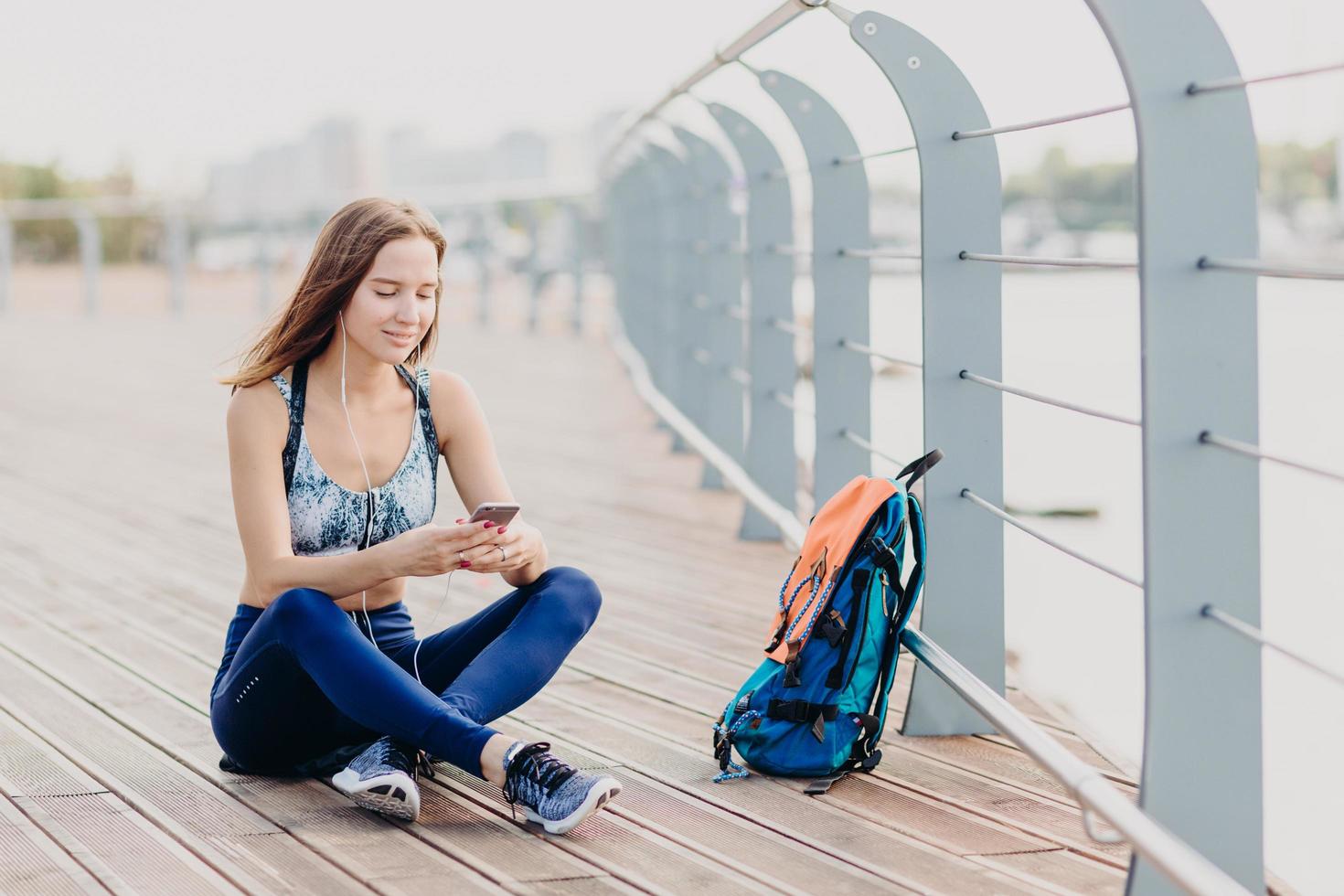 foto de una encantadora mujer satisfecha que le gusta el deporte y el estilo de vida activo, usa top y calzas, lee noticias positivas en internet en el celular, descansa después de recorrer largas distancias a pie, escucha música