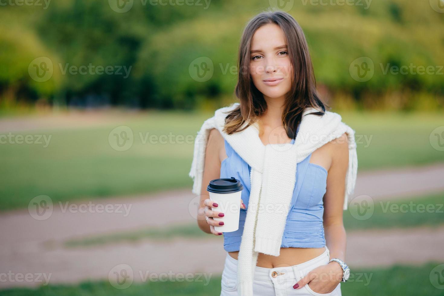 Outdoor shot of brunette woman dressed in casual t shirt, white trousers, keeps hand in pocket, drinks aromatic coffee during morning walk, stands against green nature background. Leisure concept photo