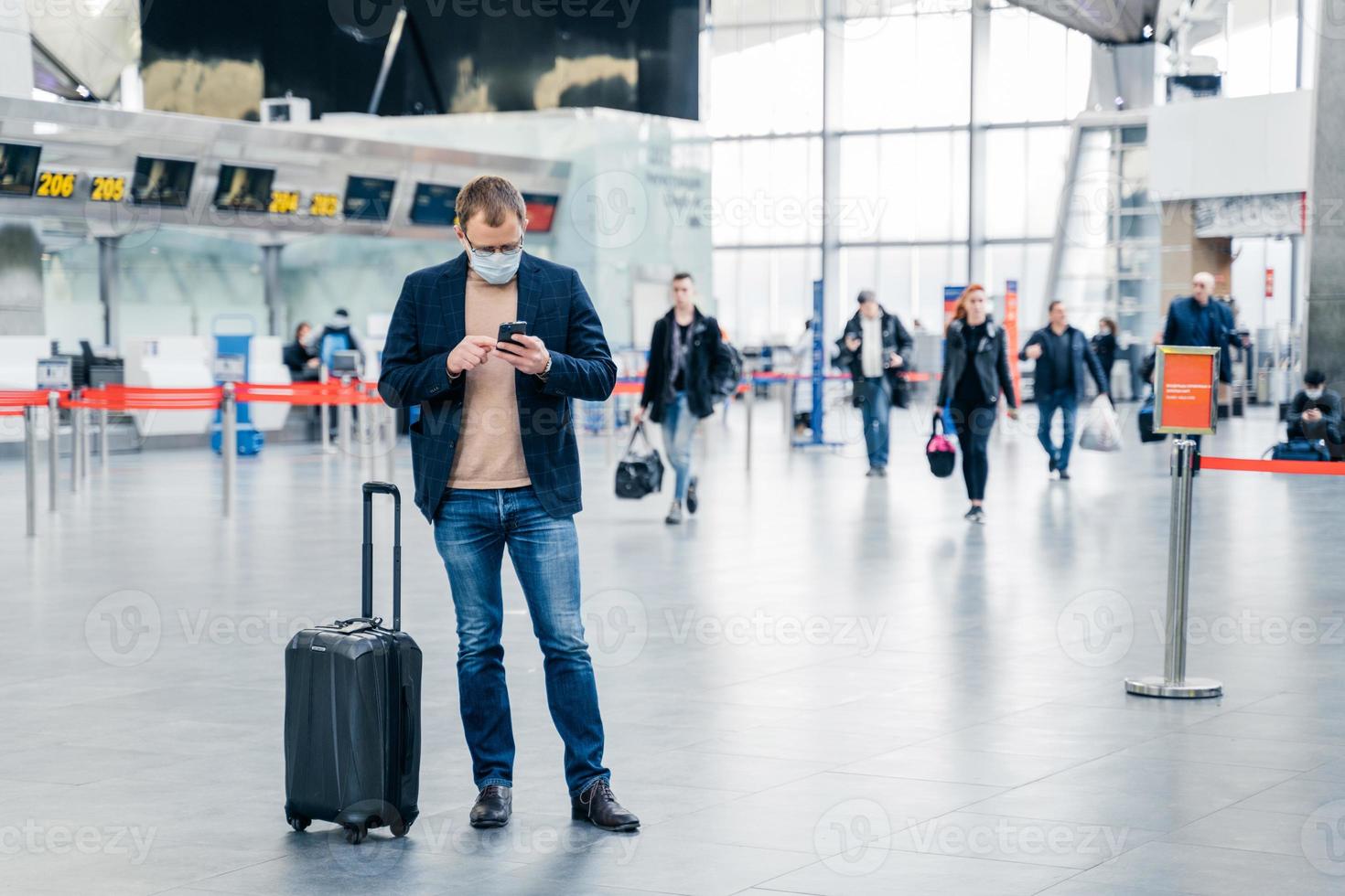 Horizontal shot of man poses in crowded airport, uses cell phone, checks time of flight online, stands near suitcase, wears medical mask during coronavirus crisis. Health care, traveling concept photo