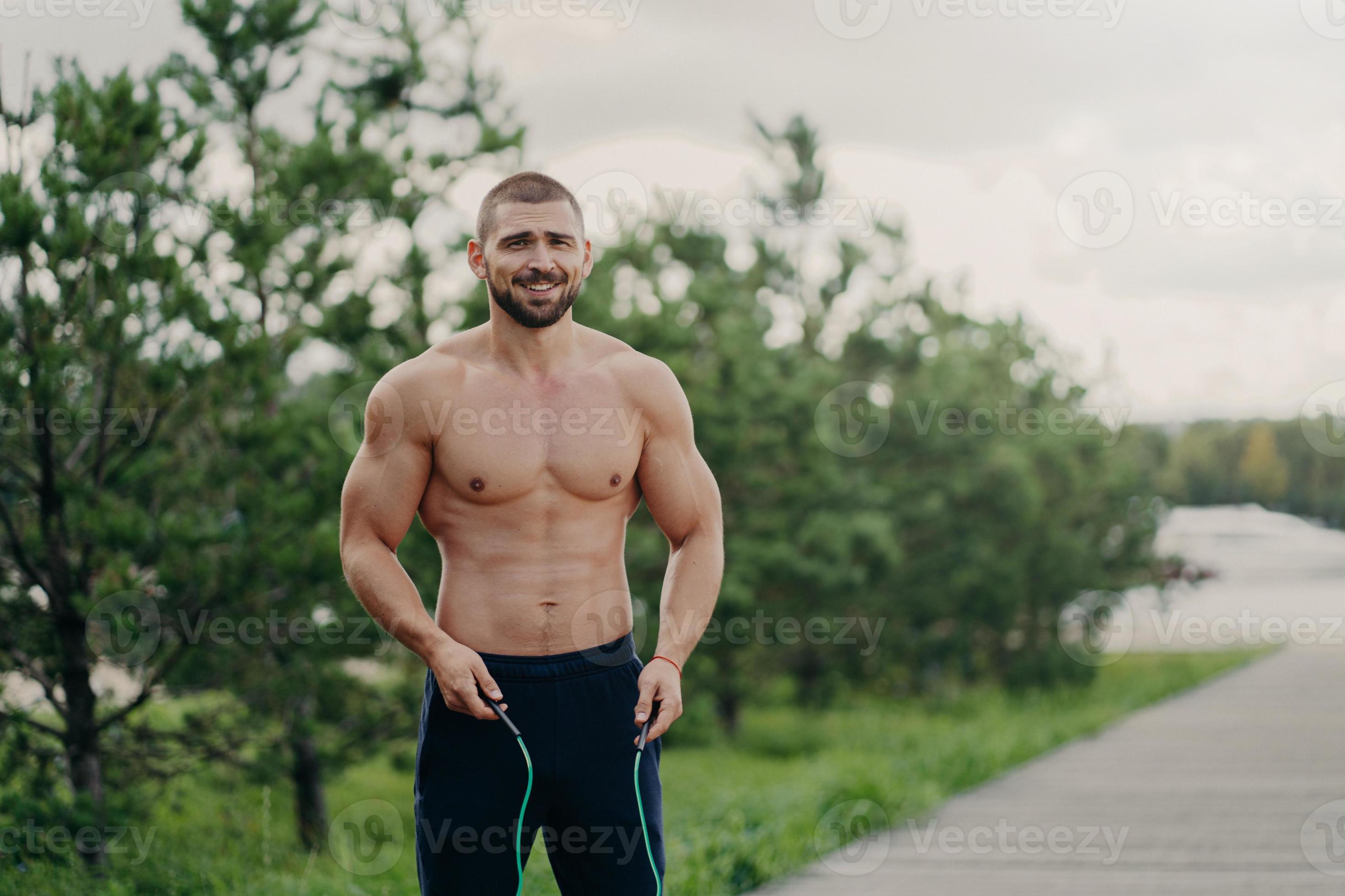 Cheerful unshaven sportsman skips with rope, has muscular body, enjoys  fitness training, poses outdoor. Strong man with naked torso uses sport  equipment for workout, breathes fresh air in nature 8067656 Stock Photo