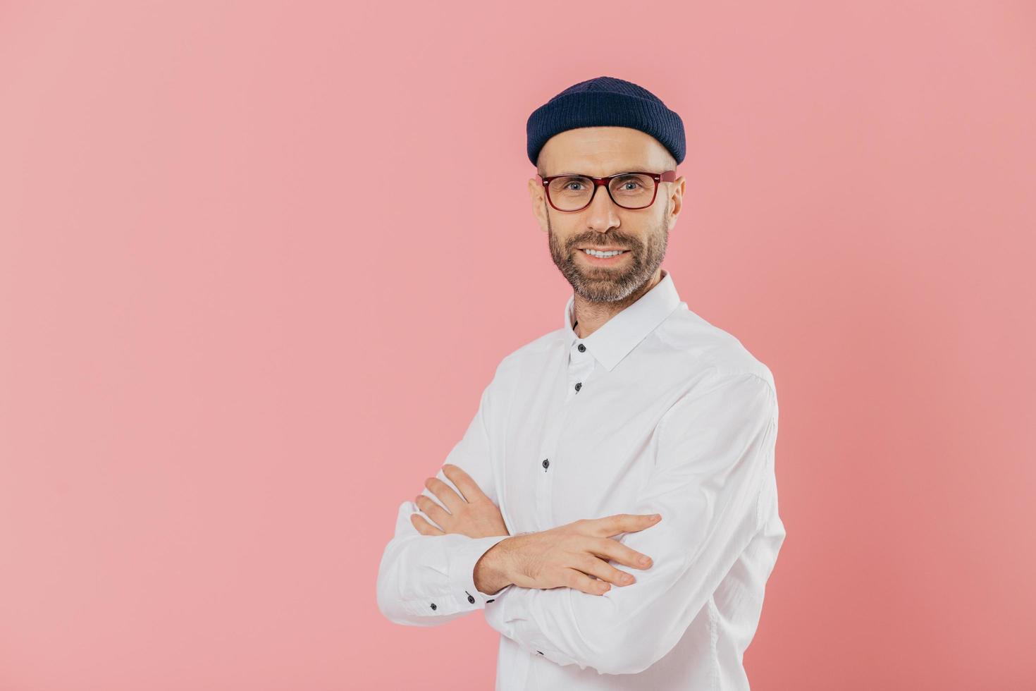 Positive male entrepreneur with self confident satisfied facial expression, keeps hands crossed, stands in profile, looks directly at camera, poses indoor against pink background with free space photo