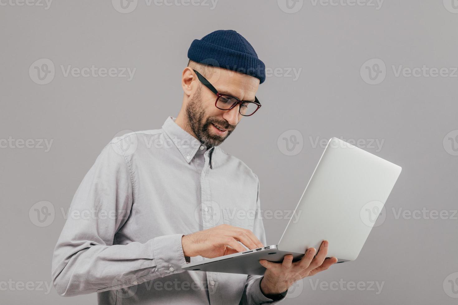 Concentrated bearded male administrative worker coordinates work on distance, searches information on laptop computer, wears white formal shirt and transparent glasses, poses indoor over grey wall photo
