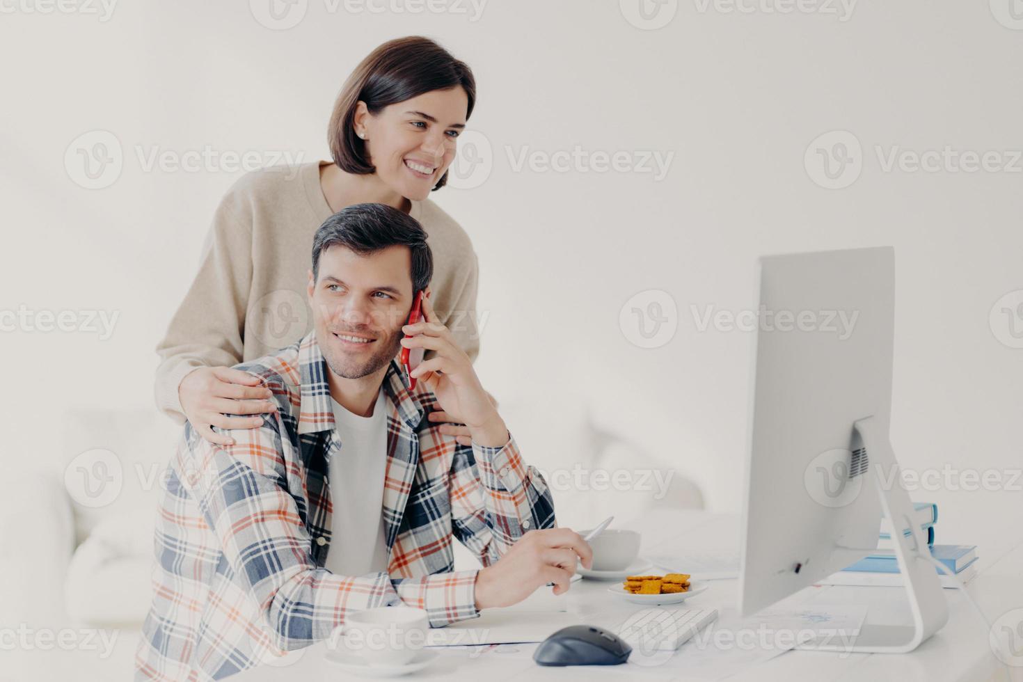 Happy young man holds mobile phone, discusses issues, manages home accounts with wife, pose in coworking space, sit in front of computer, discuss family expenses. Couple collaborate together photo