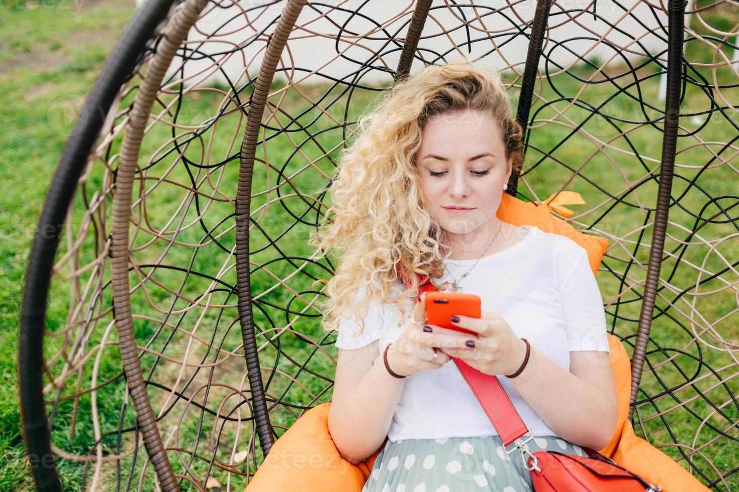 Horizontal shot of pretty young female rests in hanging chair in garden, uses smart phone, being always in touch, checks her email box, dressed casually, has good rest outdoor. Technology concept photo